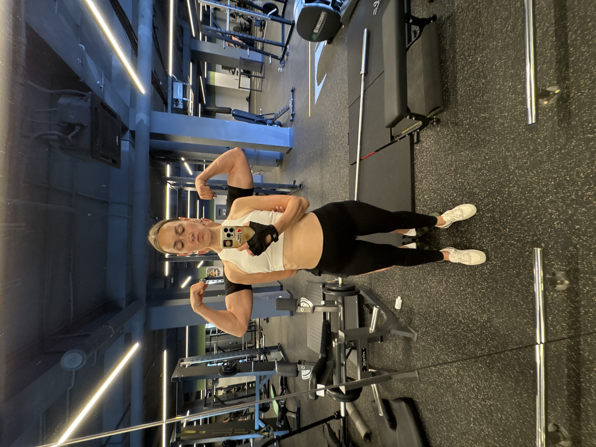 The media file shows a person taking a mirror selfie in a gym. The subject, a woman with blonde hair tied back, is wearing a white crop top, black leggings, and white sneakers. She is flexing her biceps, and her expression appears determined. She is also wearing black workout gloves.
The gym is well-lit with bright, strip lighting on the ceiling. Various gym equipment, including weight racks, benches, and weightlifting bars, are visible in the background, some of which are reflected in the large mirror in front of the subject. The floor is a speckled dark grey material commonly found in gyms. The overall atmosphere suggests an active workout session. The reflection in the mirror shows the subject and the gym environment from a different angle.
The scene is set indoors, in a modern gym. There are no indications of the time of day or weather conditions. The location context provided is Sochi, Russia, though there are no specific visual cues in the image to confirm this. No discernible text is visible in the image, apart from what might be on the phone case.