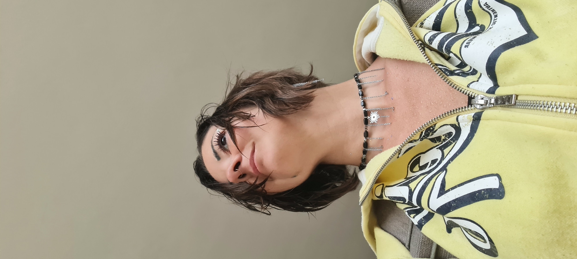 A vertical, medium-shot photograph shows a person with short, brown, wet hair and dark eyebrows turned toward the right side of the frame, wearing a black beaded necklace with small silver chains and a yellow and white hoodie with the zipper partially open. The background is a neutral light brown or tan color. The person has fair skin, full lips, and is wearing eye makeup. They also have a nose ring. The hoodie has a design in white and black on the right side. The photograph's location cannot be determined from the visual information provided.