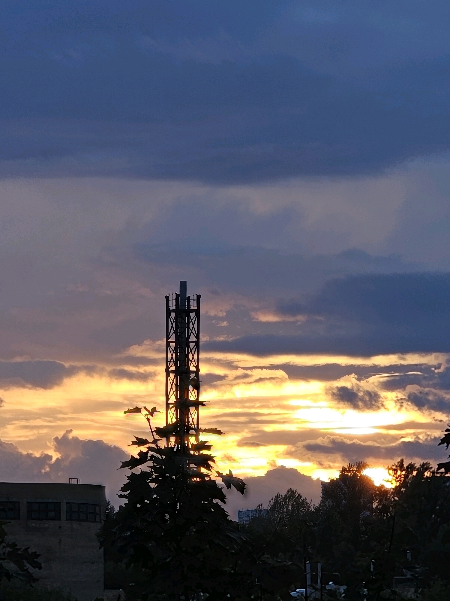 This is a low-angle shot of a radio tower and buildings against a dramatic sunset sky. The sky is filled with dark, stormy clouds mixed with bright bands of orange and yellow sunlight. A dark, silhouetted tree with large leaves is in the foreground, partially obscuring a large, grey, industrial-looking building with rectangular windows. A tall, lattice radio tower stands to the right of the building, also silhouetted against the vibrant sky. In the distance, more trees and buildings are visible, with hints of more construction. The overall impression is one of an urban landscape at dusk, with the natural beauty of the sunset contrasting with the man-made structures.