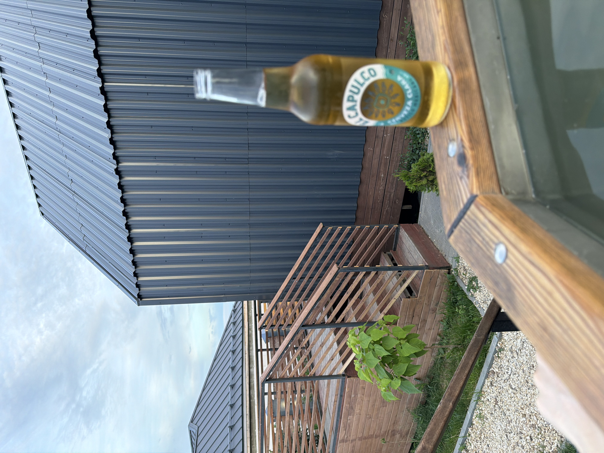 An eye-level, medium shot captures a bottle of Acapulco Cerveza Clara beer resting on the wooden edge of what appears to be a hot tub. The beer bottle is positioned in the foreground, slightly out of focus, with its label visible. Behind the bottle, a dark gray, corrugated metal building dominates the background, with a contrasting lighter gray metal roof visible in the upper left portion of the frame. To the left of the building, a wooden deck and staircase with railings are visible, leading to a lower level with gravel and some greenery. The sky above is overcast with scattered clouds. The scene suggests an outdoor relaxation area, possibly at a residential property or a small resort. The specific location within Семилуки, Russia, is not discernible from the visual information alone.