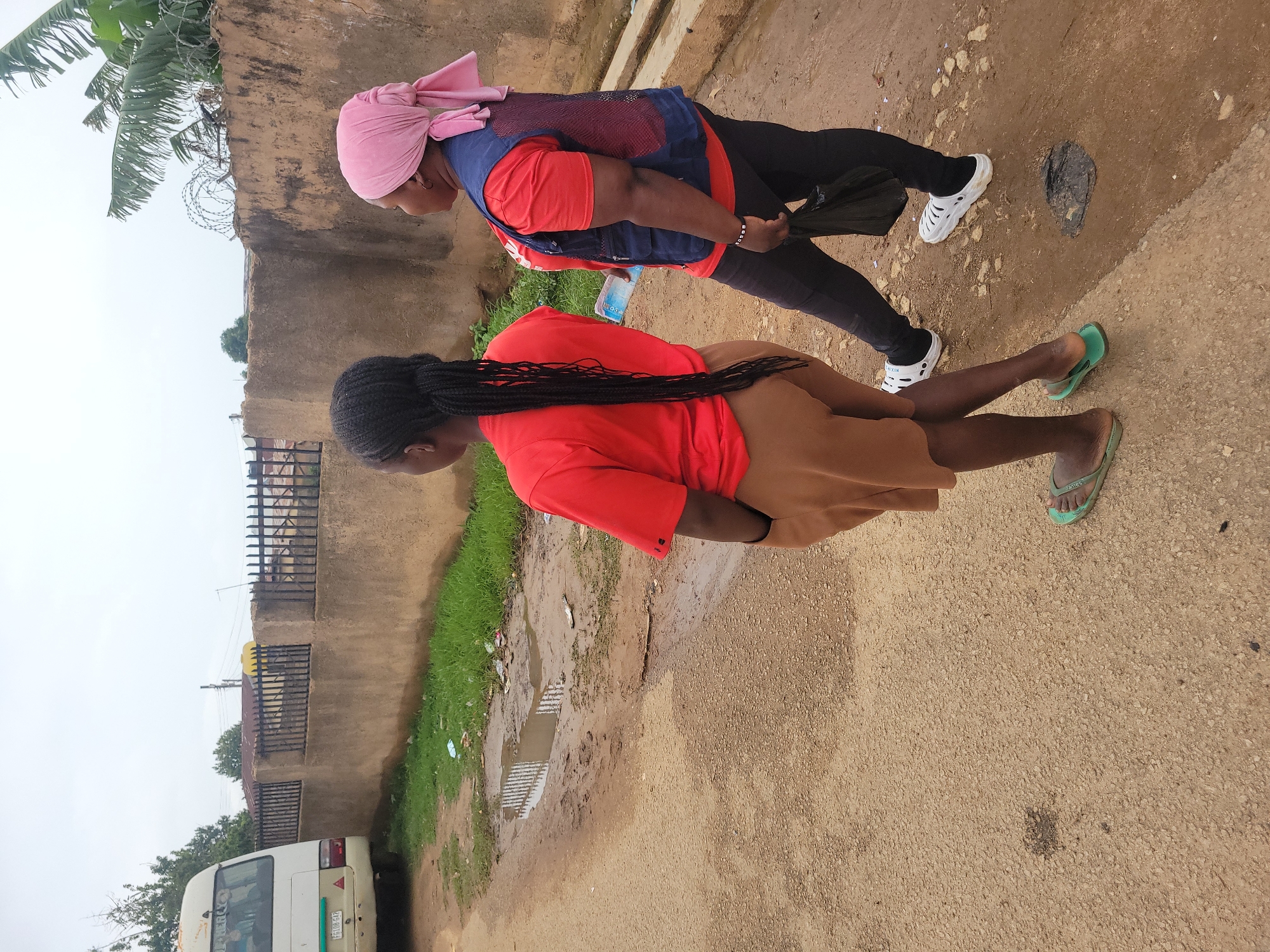 Two women are walking side-by-side on a dirt road in Jos, Nigeria. The woman on the left is wearing a red t-shirt and tan shorts, with her hair in braids. She is wearing green flip-flops. The woman on the right is wearing a red t-shirt, a blue vest, and black leggings. She has a pink headscarf on and is wearing white clogs. She is holding a black plastic bag in her right hand and a water bottle in her left. A white bus is parked on the left side of the road, and a low concrete wall with a metal fence runs along the right side. There is some grass and debris along the side of the road, and a puddle of water in the road. The sky is overcast.