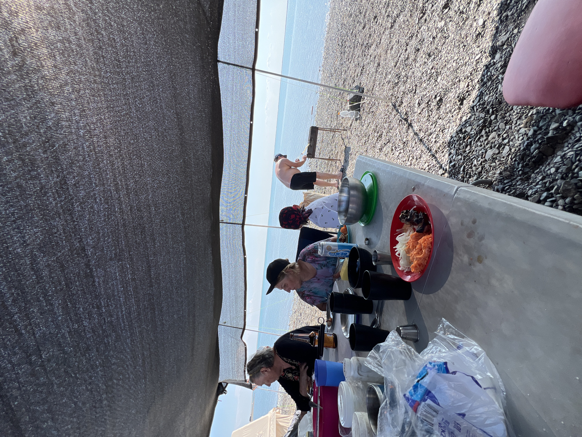 An outdoor barbecue is taking place on a rocky beach, presumably in the city of Karashygan, Kazakhstan, with the Caspian Sea visible in the background. A group of four people is present. Two women are seated at a table under a dark mesh canopy, one with a bottle of beer and the other with a can of what appears to be Beks Energy Drink. A child wearing a red and white bandana is also seated at the table. A man, shirtless and wearing black shorts and a cap, is standing and grilling food on a barbecue. On the table, there are various items including plastic cups, metal bowls, and a red plate with food. A portion of a pink object, possibly a seating cushion, is visible in the lower right corner of the image. The environment consists of a pebble beach and calm blue water under a clear sky.