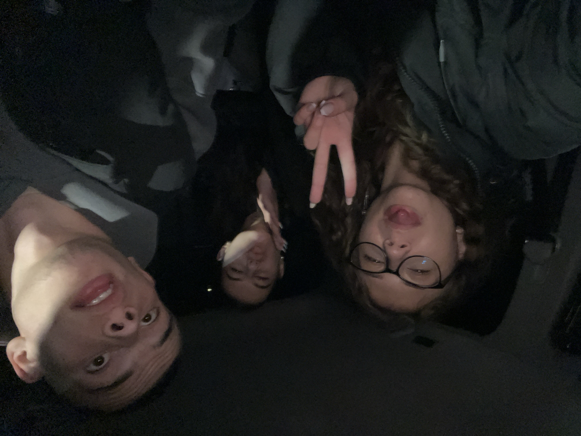 Three individuals are present in the image. On the left, a male subject with short hair is looking up. In the center, another individual is visible, with only the face and upper body visible. On the right, a female subject with curly hair and glasses is sticking out her tongue and making a peace sign with her fingers. The subjects appear to be in a dimly lit environment, potentially a car interior. There are no visible landmarks or details to confirm the location as Karavelovo, Bulgaria.
