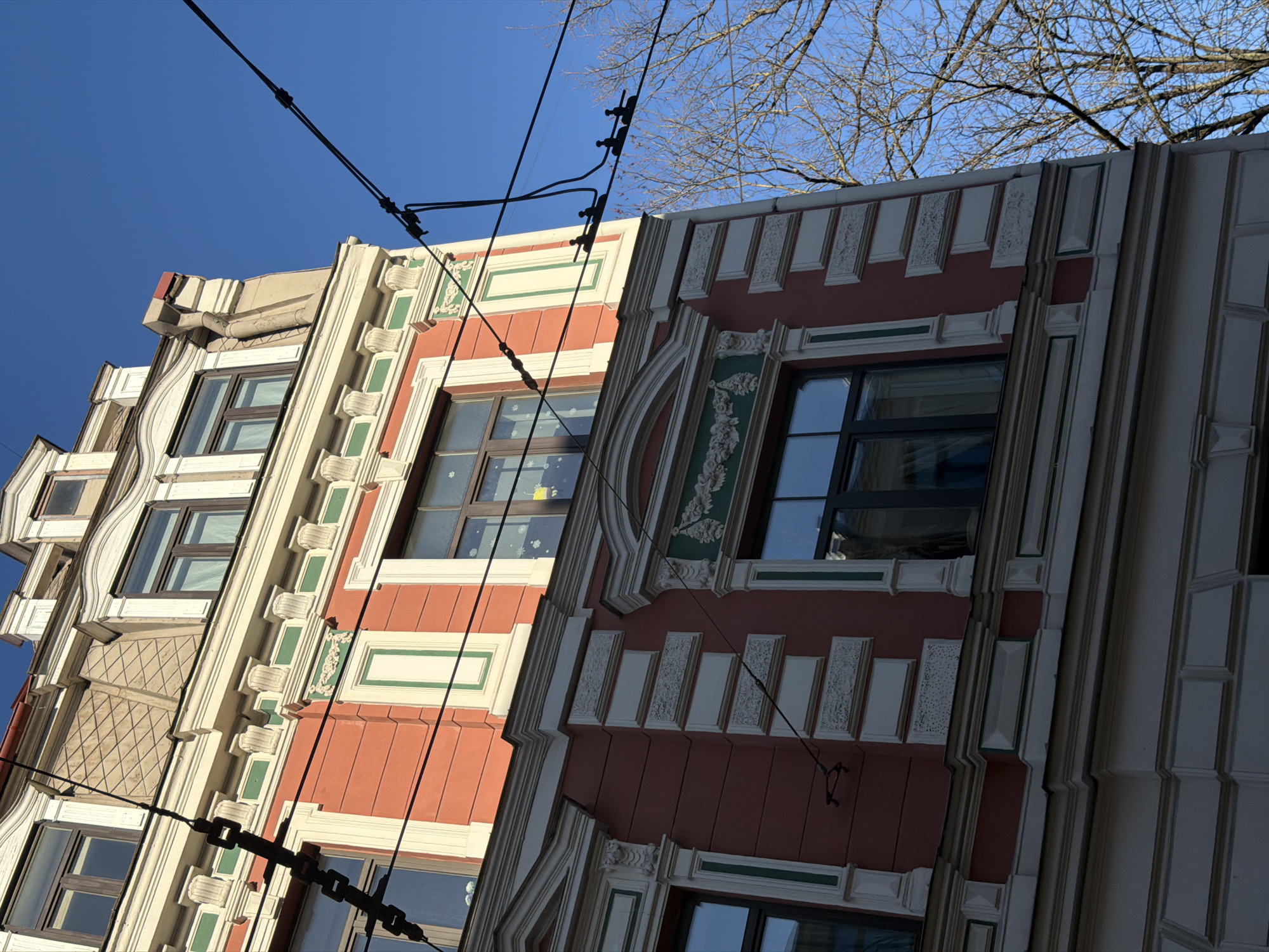 The image shows two colorful buildings in Riga, Latvia, viewed from a low angle. The building on the left is orange and cream with green accents, and the building on the right is a darker red with cream and green detailing. Both buildings feature decorative windows and architectural details. Overhead power lines are visible against the clear blue sky. Bare tree branches appear in the upper right corner of the frame.