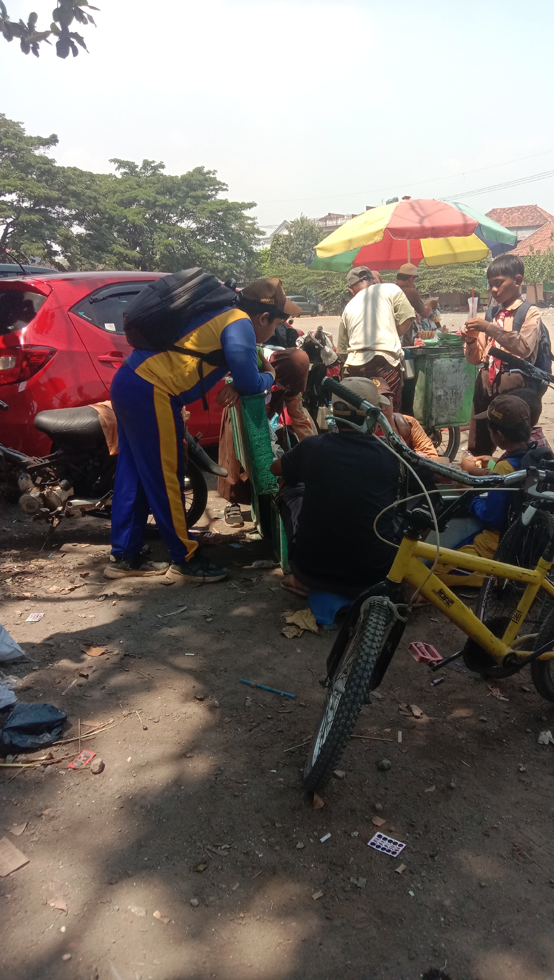 The image depicts a street scene in Ngempit, Indonesia, on a sunny day. Several people, some appearing to be schoolchildren in uniform, are gathered around a cart selling drinks. The cart has a colorful umbrella providing shade. In the foreground, a yellow bicycle is parked. To the left, a red car and a motorcycle are visible. The ground is dusty and uneven, with scattered debris. The atmosphere suggests a casual, everyday moment in a local community.