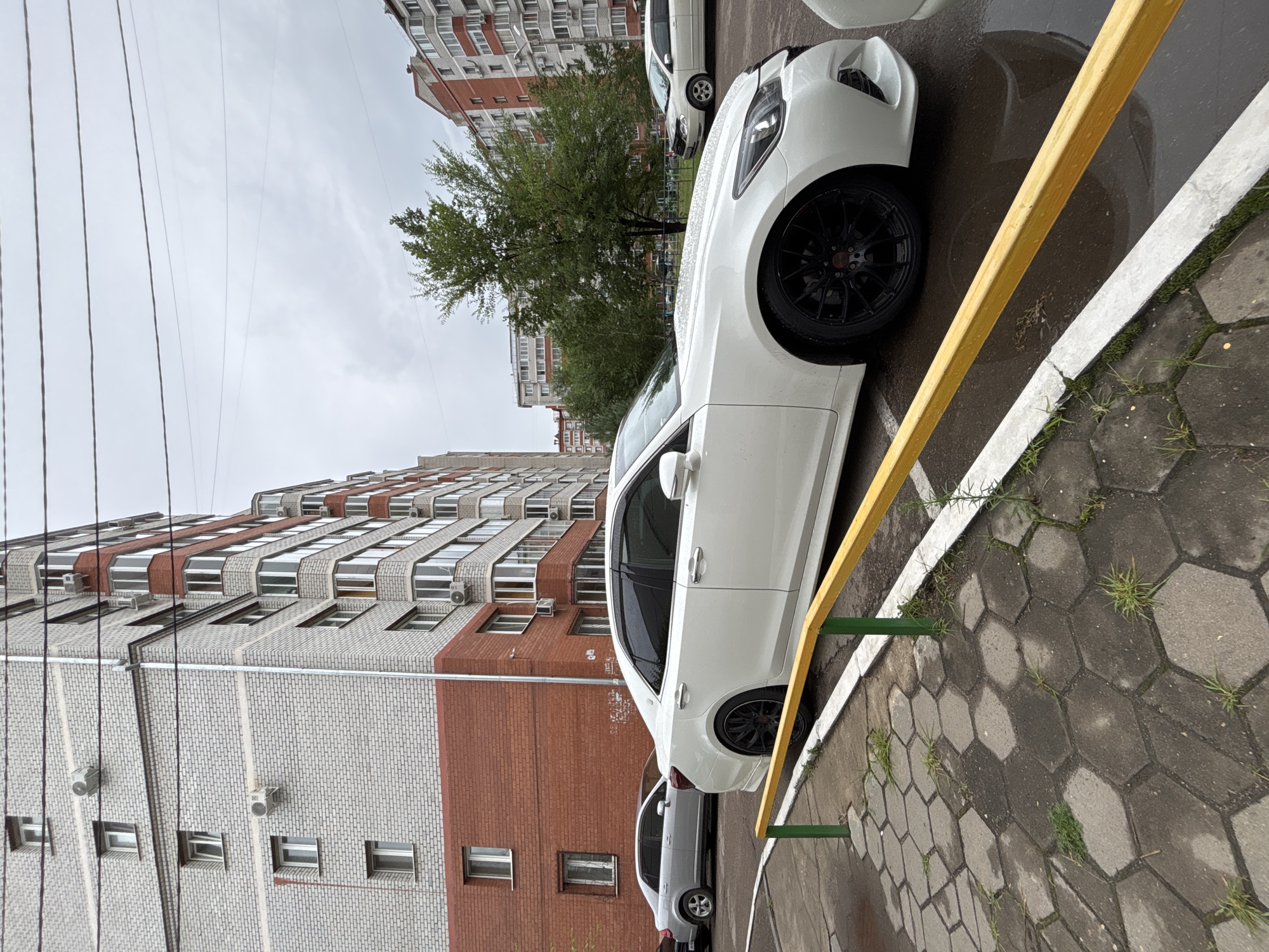This is a medium shot, eye-level, outdoor photograph taken on a cloudy day in Blagoveshchensk, Russia. The foreground features a wet, hexagonal-patterned concrete walkway with sparse grass growth. A yellow metal barrier with green posts separates the walkway from a paved area where a white sedan is parked. The car is facing to the right, with its rear wheel and part of its body visible. Behind the car, a light-colored sedan is parked. In the background, a tall, multi-story brick apartment building dominates the left side of the frame, characterized by its red brick base and lighter brick upper floors, with numerous windows and air conditioning units. To the right of the building, a large, leafy green tree stands. Further in the background, another apartment building with red and white facades is visible. Power lines are present in the upper portion of the sky. The overall environment suggests a residential urban setting.