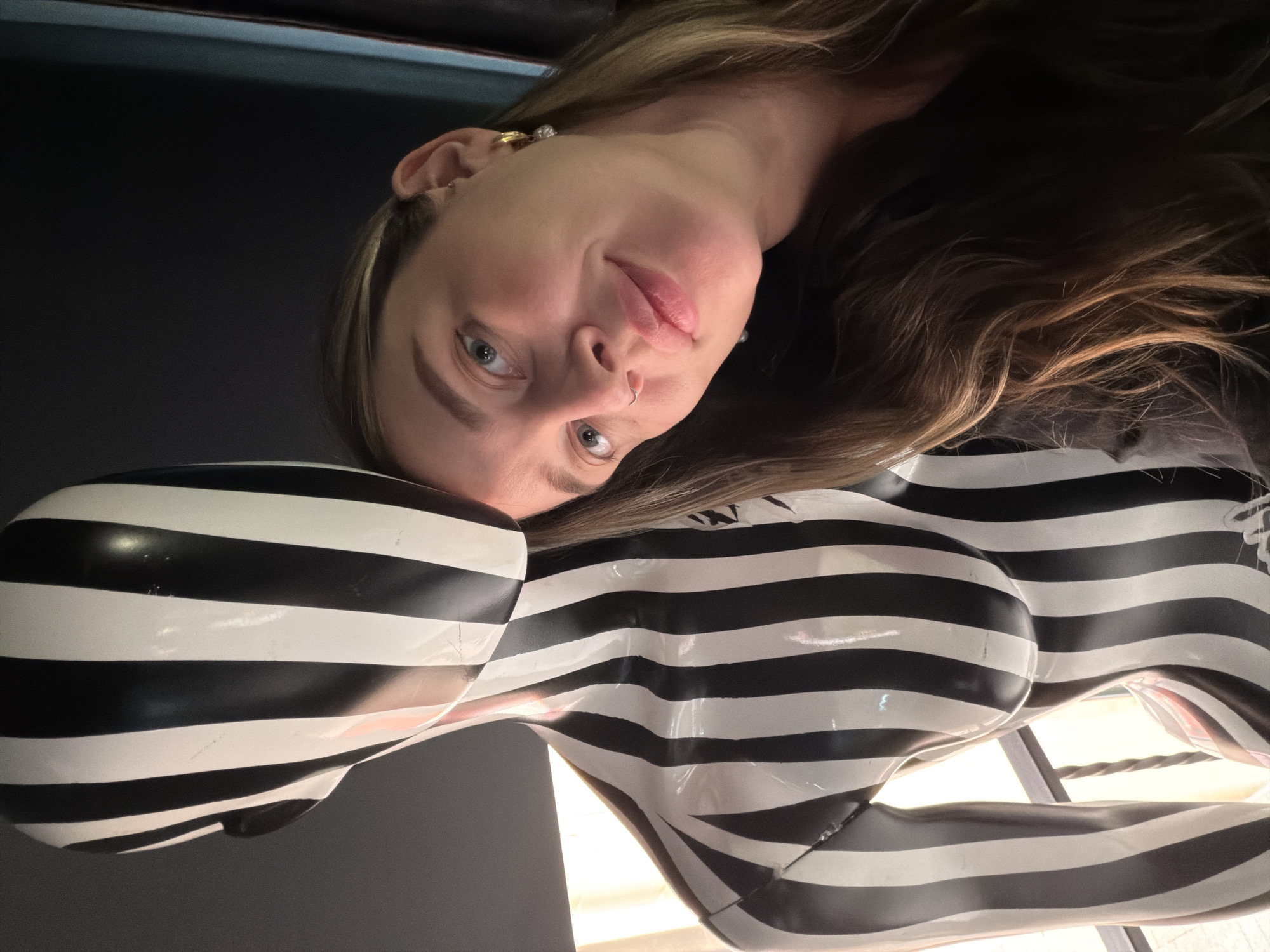 The media file displays a portrait-style photograph, rotated 90 degrees counter-clockwise, featuring a young woman and a distinct art sculpture in an indoor setting. The woman, with long, wavy light brown hair and subtle blonde highlights, has light blue eyes and a small silver hoop nose piercing. She wears small, pearl-like earrings and looks directly at the viewer with a calm, soft smile. Her head is positioned to the left, resting gently against a unique, glossy sculpture that dominates the foreground. This sculpture is a stylized female torso and hip section, appearing headless, armless, and legless, entirely covered in bold, horizontal black and white stripes. Light reflects off its polished surface, indicating a well-lit but possibly dark-walled environment. The background is dark and unfocused, suggesting an art gallery or a similar exhibition space. No text is visible in the image. The scene is located in Moscow, Russia.
