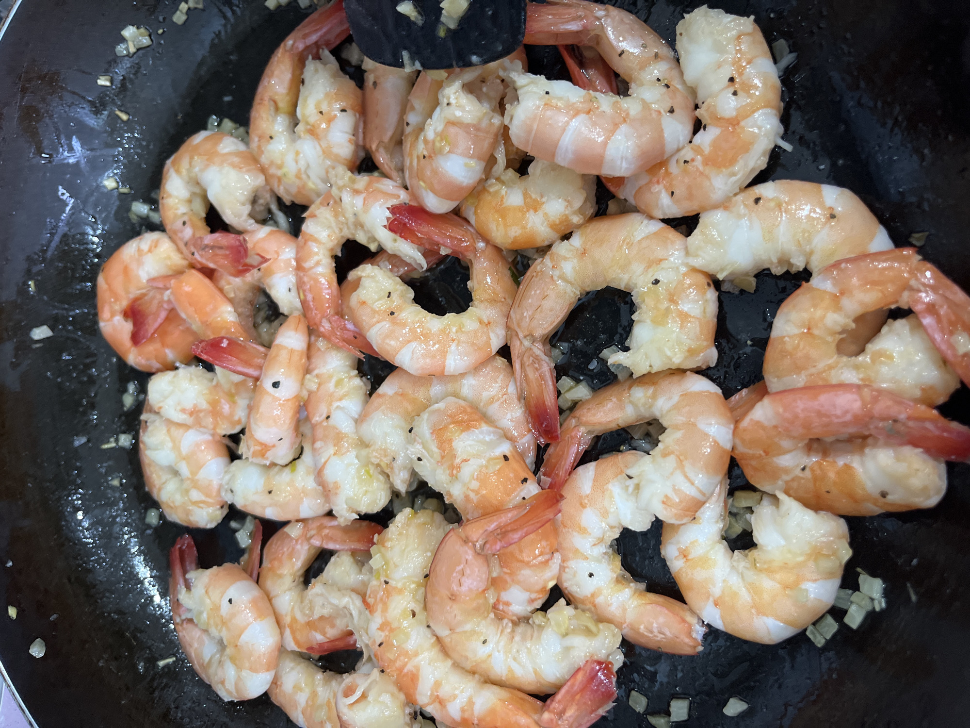 The image shows a close-up of cooked shrimp in a dark-colored pan, likely a wok or skillet. The shrimp are pink and white, seasoned with black pepper and interspersed with small pieces of what appears to be minced garlic. The environment seems to be a kitchen setting. There is no explicit evidence to suggest the location is specifically in the Barrio de Nuevo León, Mexico. No people are visible.
