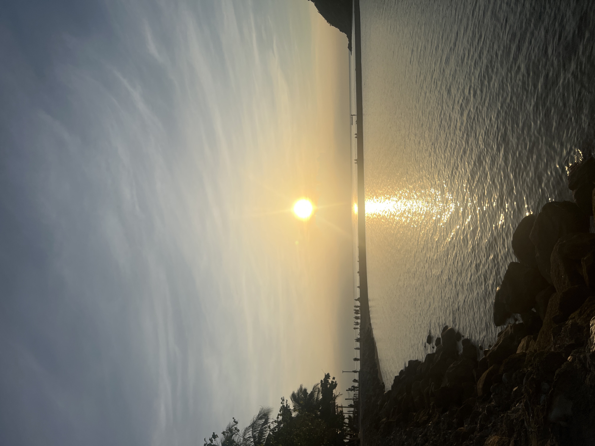 The image depicts a sunset over the sea in Ko Pha-ngan District, Thailand. The sun, a bright orb with a lens flare, is low in the sky, casting a golden glow across the water. The sea is calm, with gentle ripples reflecting the sunlight. In the foreground, a rocky shoreline is visible, with lush green trees lining the edge of the land. A group of people can be seen walking along the shore, silhouetted against the setting sun. In the distance, a small island or landmass is visible on the right side of the frame. The sky is a pale blue with wispy clouds, suggesting the end of the day. The overall atmosphere is serene and tranquil.