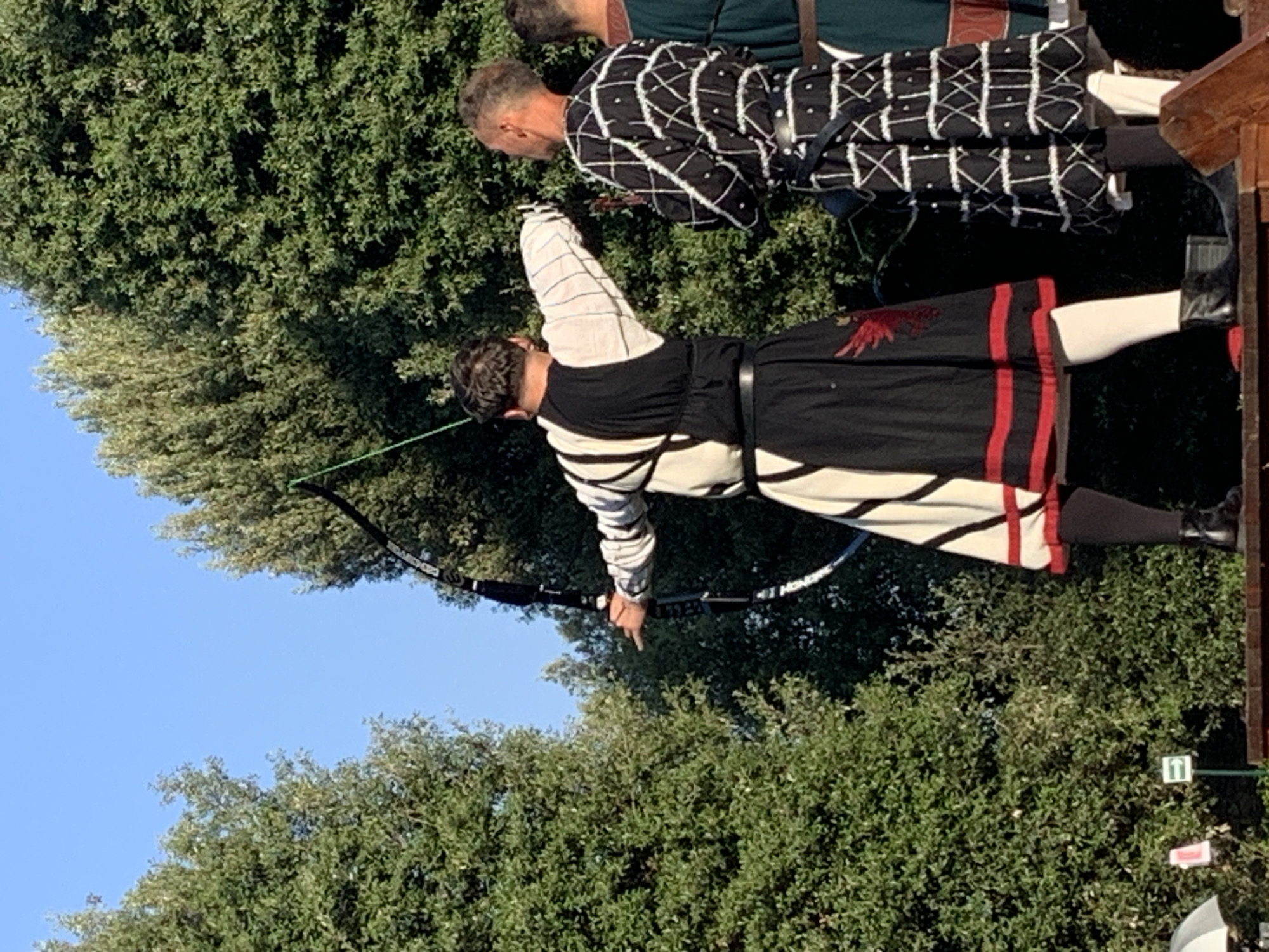 A reenactment of historical archery is taking place outdoors, possibly in San Quirico d'Orcia, Italy, given the context of the image. Three individuals, dressed in medieval-style attire, are the primary subjects. The foreground features a person aiming a bow and arrow, drawing the string back in preparation for a shot. This individual wears a white, long-sleeved tunic with black and white striped sleeves, a black vest, and a black skirt with red trim and a red embroidered emblem. They are also wearing white stockings and black boots. To their right stands another person in a black tunic with a white geometric pattern, black belt, white stockings, and black shoes. Further to the right, a third individual is partially visible, wearing a green tunic with a brown belt. The background is dominated by lush green trees and a clear blue sky. The individuals are standing on a raised wooden platform, suggesting a performance or event setting.