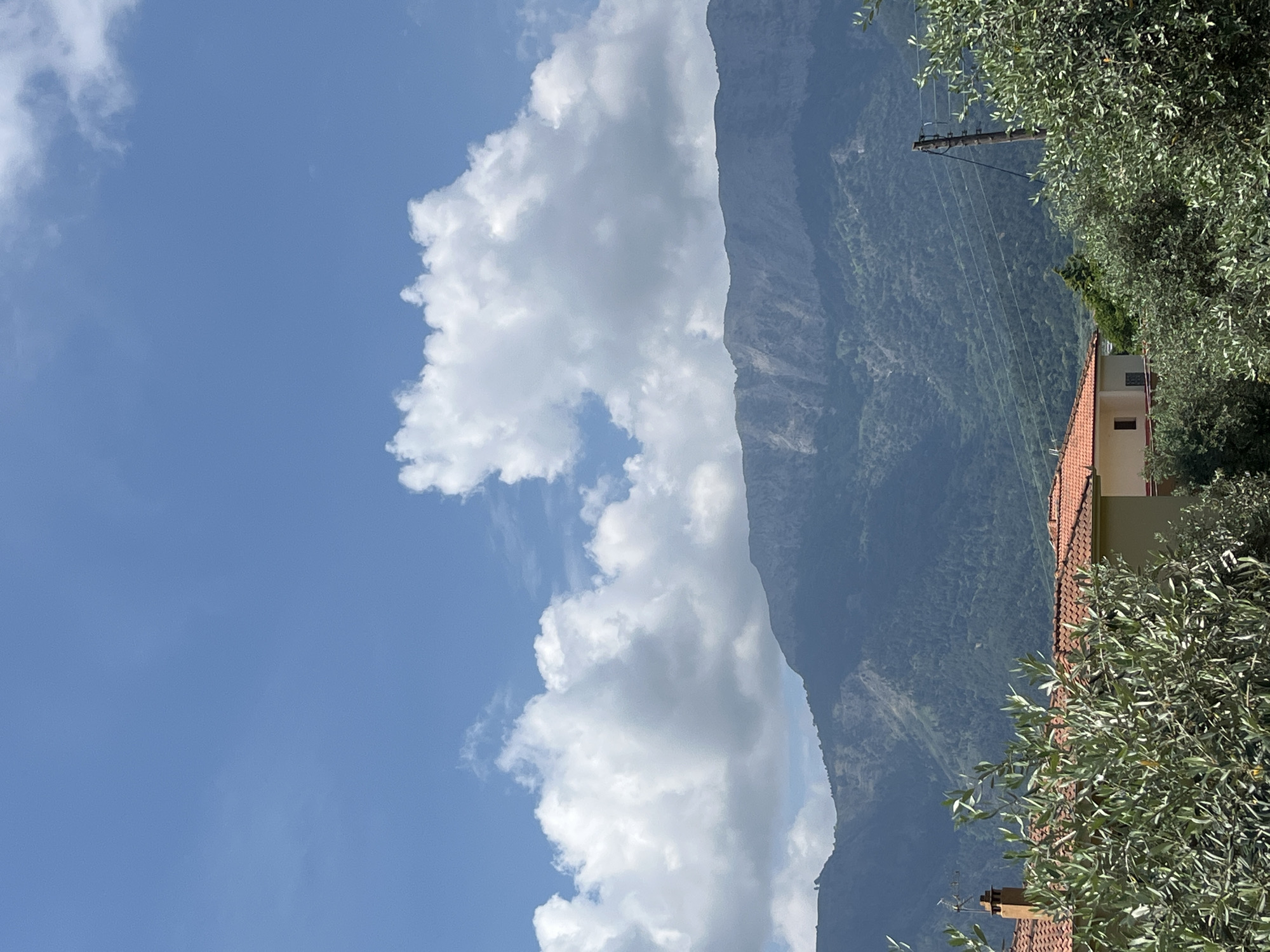 The image shows a view of a mountainous landscape with a residential building and vegetation in the foreground. The sky is blue with scattered white clouds. In the middle ground, a range of mountains is visible, with some vegetation and houses on the slopes. In the foreground, there is a house with an orange tiled roof and a chimney. Olive trees with green leaves are in front of the house. A utility pole with wires is also visible. This scene is located in Golden Beach, Greece.