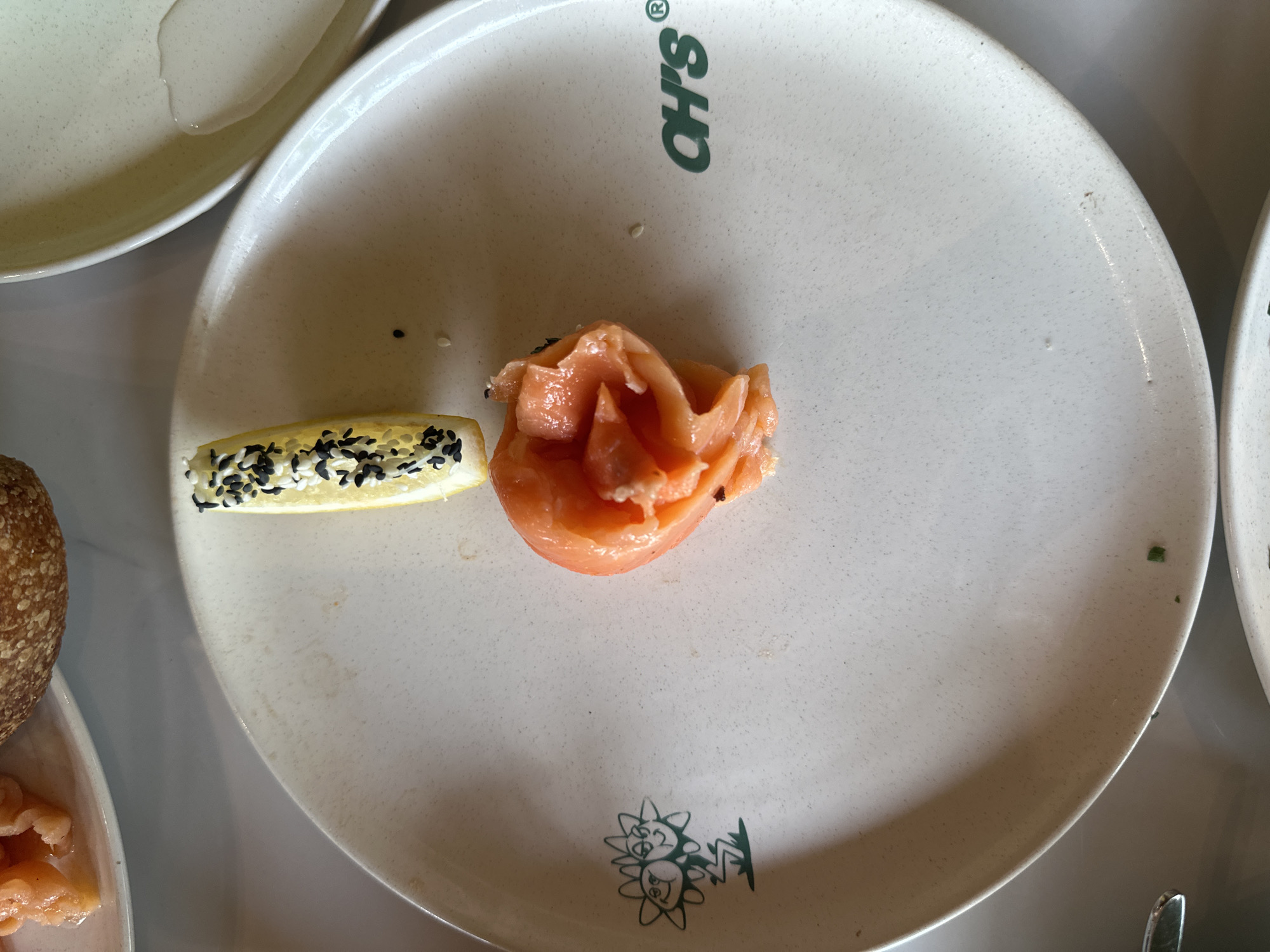 The image displays a close-up, overhead view of a single serving of food on a light-colored, speckled plate. The main subject is a delicate portion of raw fish, likely salmon, arranged in a rose-like shape, with a soft orange-pink hue. Beside it, to the left, is a wedge of lemon, generously topped with a mix of black and white sesame seeds. A few loose sesame seeds are scattered across the plate.
The plate itself features distinctive green branding: the text "OH'S®" is visible at the top, and a playful cartoon-like illustration of two sun or flower figures is positioned towards the bottom. The plate's surface has a subtle, textured appearance.
The scene suggests a dining context, possibly in a restaurant in Mengwi, Indonesia, where this particular dish is being served. Portions of other plates are visible in the periphery: one to the upper left appears to contain some liquid, while another to the lower left shows part of a brown, bun-like food item and what looks like more of the same raw fish. The lighting is even, suggesting an indoor setting. The focus is entirely on the food presentation, with no people visible.