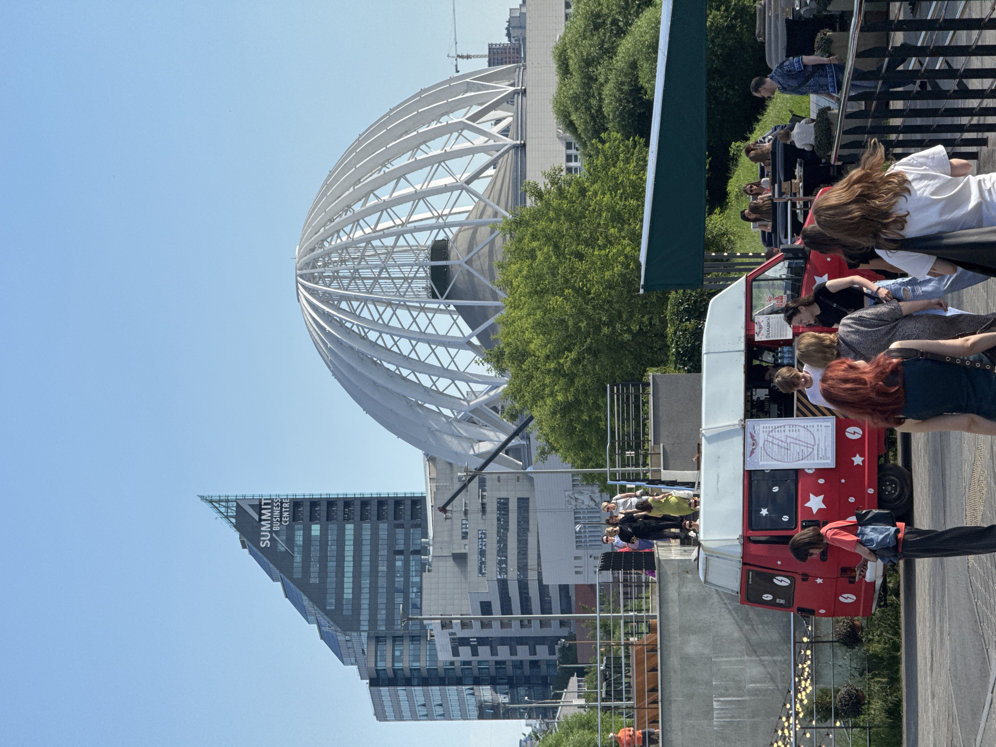 The image depicts an outdoor scene with a red food truck and a group of people in Yekaterinburg, Russia. The food truck, decorated with white stars and coffee beans, appears to be a central point of activity. Several individuals are gathered around it, suggesting a purchase or social interaction. Some are standing, while others are seated or leaning.
In the background, a distinctive geodesic dome structure with a white lattice framework dominates the sky. Adjacent to it, a modern, angular skyscraper with the words "SUMMIT BUSINESS CENTRE" is visible, providing a clear location cue. Lush green trees frame the scene, suggesting a pleasant outdoor environment. The sky is a clear, bright blue, indicating a sunny day, likely midday due to the strong sunlight.
People are dressed in casual summer clothing, and their postures suggest a relaxed atmosphere. Some are looking at their phones, while others are engaged in conversation. The overall mood seems to be one of everyday life in an urban setting, with the food truck serving as a hub for community interaction. The image captures a blend of modern architecture, natural elements, and human activity.