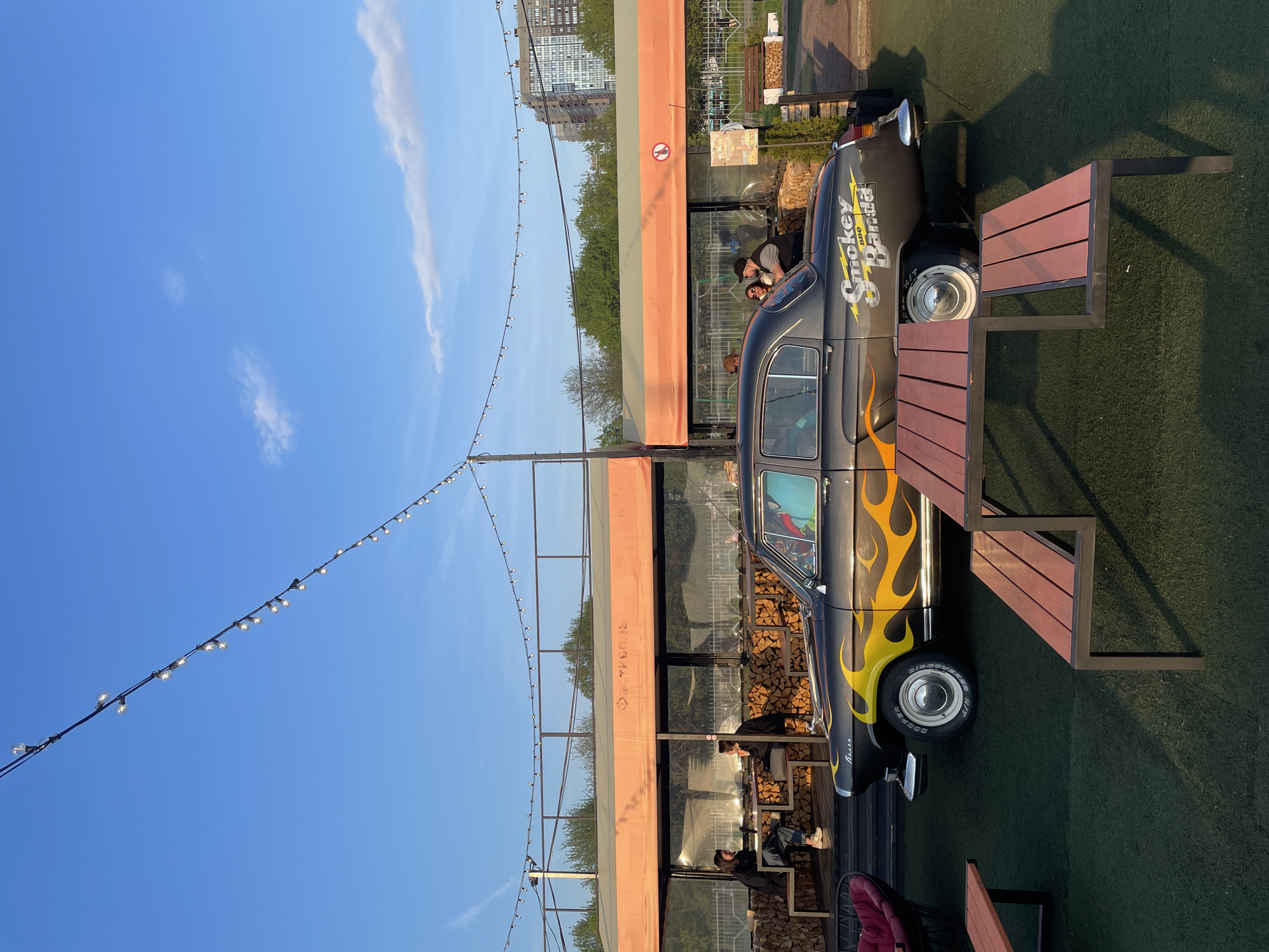 The image shows a modified vintage car with a "Smokey Panda" logo and flame decals parked next to a table with benches, adjacent to an outdoor structure with wooden stacks inside. People are visible inside the structure, possibly patrons. Overhead string lights stretch across the scene against a blue sky. The setting appears to be in Saint Petersburg, Russia, suggested by the architecture and urban landscape in the background.