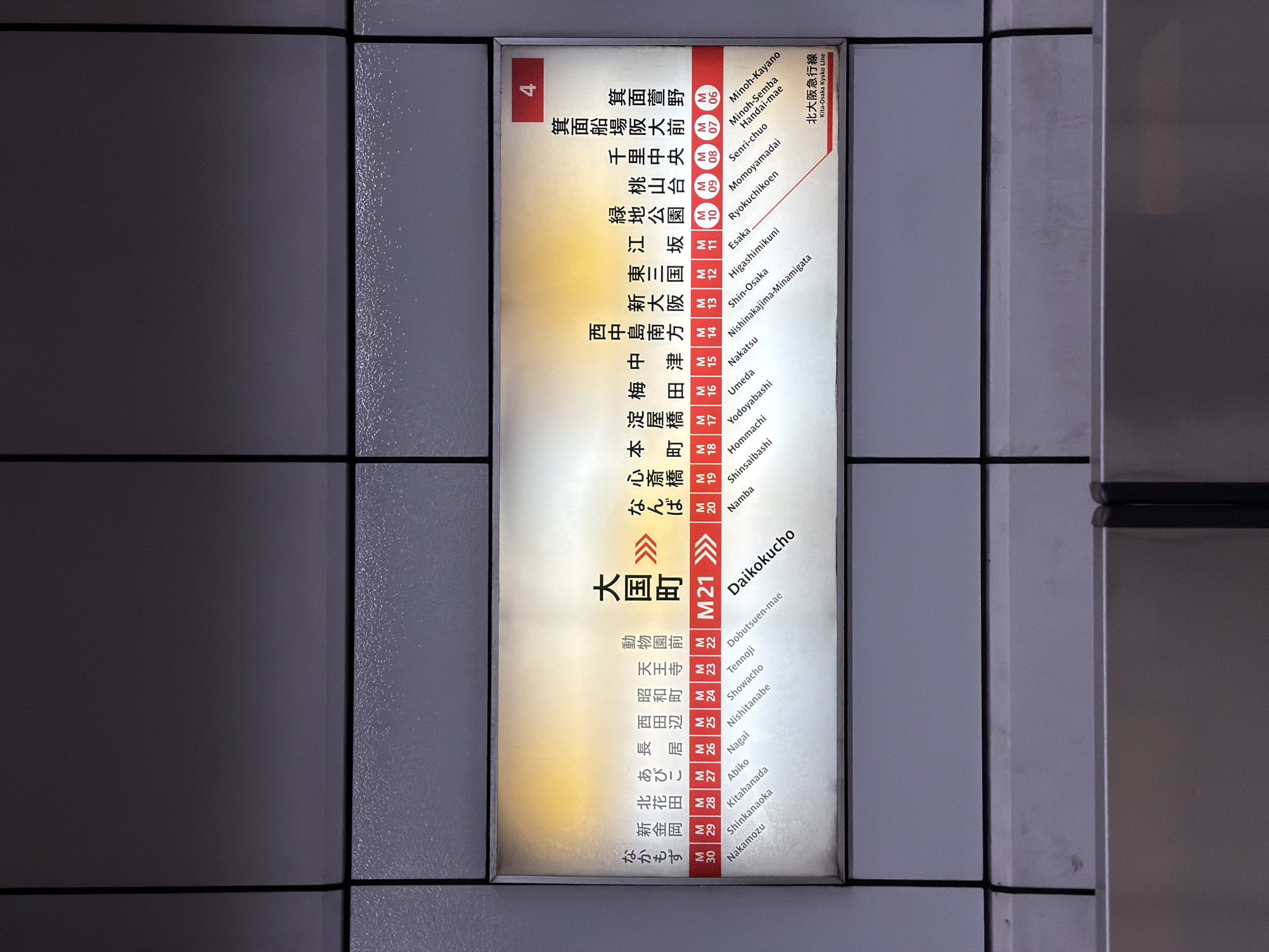 The image shows a backlit subway or train station sign in a vertical orientation, likely located within a Japanese transit station. No people are visible. The sign displays a list of station names in both Japanese (kanji and hiragana) and English romanization, each accompanied by an alphanumeric identifier (e.g., "M" followed by a number).
The station "Daikokucho" (大国町) is prominently centered and highlighted with the identifier "M21" and double arrows, indicating it is the current station. The sign lists stations including Minoh-Kayano (M06), Minoh-Semba Handai-mae (M07), Senri-chuo (M08), Momoyamadai (M09), Ryokuchikoen (M10), Esaka (M11), Higashimikuni (M12), Shin-Osaka (M13), Nishinakajima-Minamigata (M14), Nakatsu (M15), Umeda (M16), Yodoyabashi (M17), Hommachi (M18), Shinsaibashi (M19), Namba (M20), Daikokucho (M21), Dobutsuen-mae (M22), Tennoji (M23), Showacho (M24), Nishitanabe (M25), Nagai (M26), Abiko (M27), Kitahanada (M28), Shinkanaoka (M29), and Nakamozu (M30). The "Kita-Osaka Kyuko Line" (北大阪急行線) is also mentioned at the top right of the sign. A red box with the number "4" is visible at the top left corner. The surrounding environment appears to be a dark, paneled or tiled wall, consistent with an indoor station setting. The sign itself is illuminated, providing clear readability.