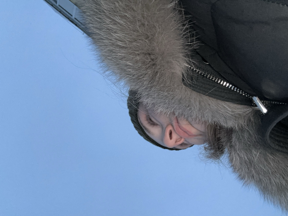 A person in a dark winter coat with a fur-lined hood faces downward. The background is a bright blue sky, with a partial building visible in the top left corner. The person wears a dark knit cap beneath the hood. A silver zipper with a pull tab is visible on the coat. The fur trim is gray and fluffy.