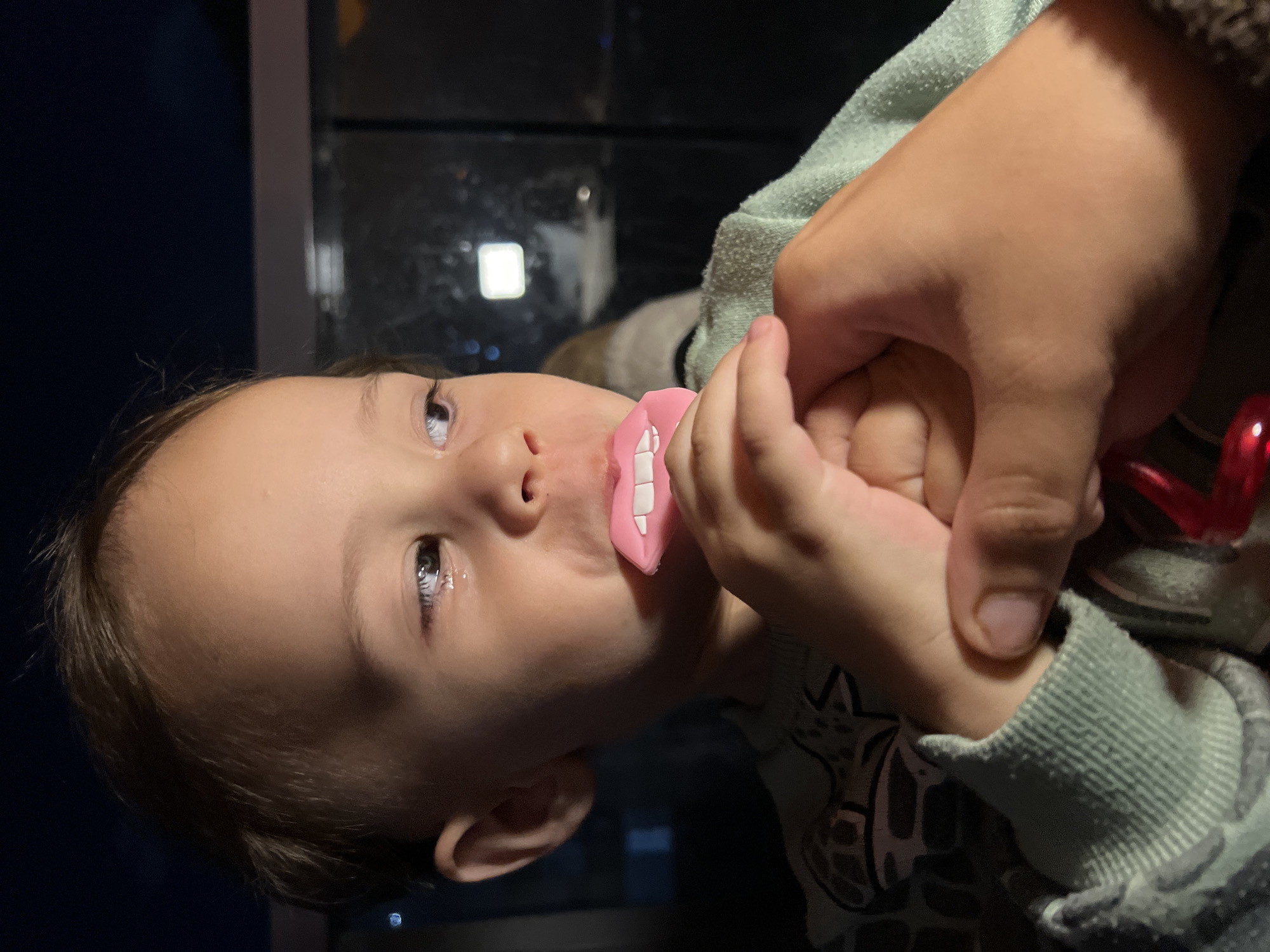 A young child is holding an adult's hand and holding a pacifier with fake teeth in their mouth. The child is wearing a green sweater. The background suggests an indoor location, possibly near a window with lights outside. There is no verifiable data to support the location of the city of Kiryat Bialik, Israel.