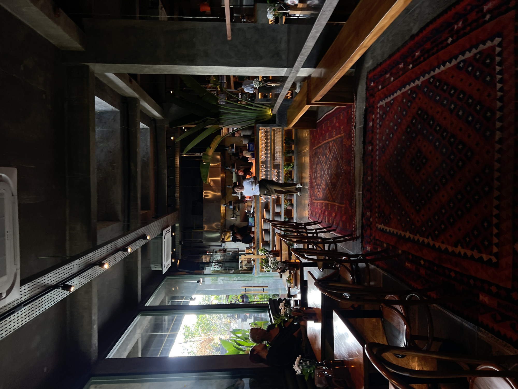 The image shows the interior of a dimly lit, modern restaurant in Mengwi, Indonesia. The restaurant features an industrial aesthetic with exposed concrete ceilings and columns, complemented by dark wooden furniture and flooring. Large windows offer a view of lush greenery outside. A long, red oriental rug is laid out on the floor, leading towards the open kitchen area.
In the foreground, several wooden tables and chairs are set for dining. Two women are seated at a table by the window, engaged in conversation. The central area of the restaurant features a long wooden bench seating area.
The open kitchen is visible in the background, with stainless steel countertops, shelves stocked with glasses and plates, and kitchen staff in uniform working. Several people are present, including staff members attending to customers or preparing food, and diners seated at tables. A woman in a white shirt and brown apron is walking across the floor carrying a tray. Another woman, also in a white shirt and brown apron, is standing near the kitchen. The overall atmosphere suggests a contemporary dining establishment with a relaxed ambiance.