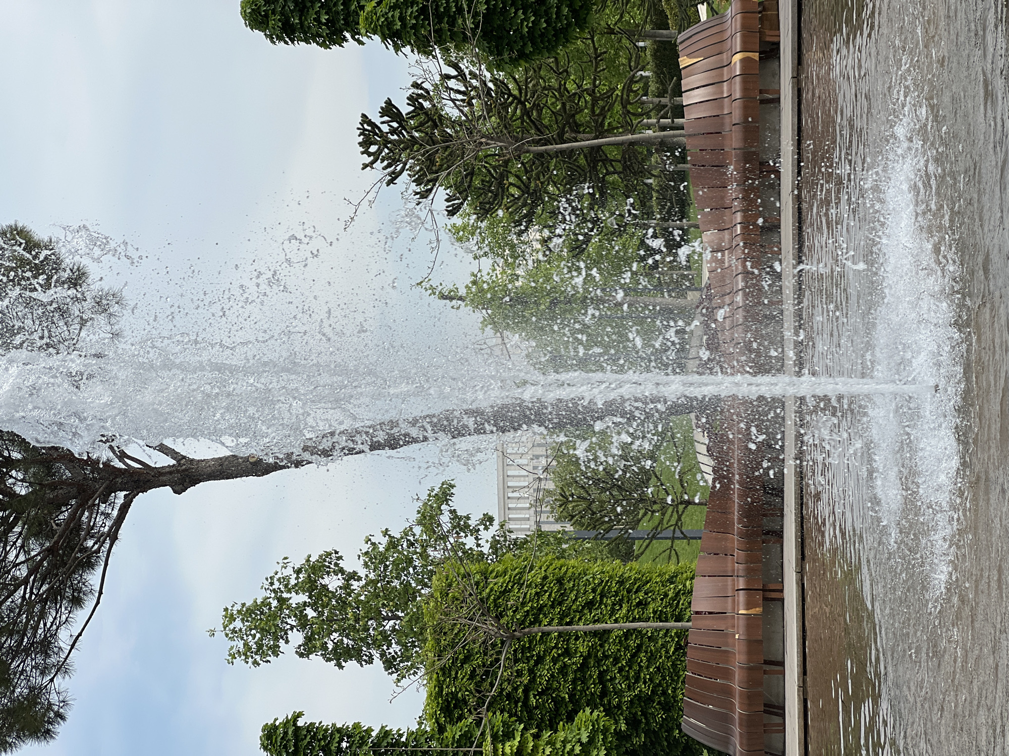 The image depicts a fountain in a park setting. Water sprays upwards in a tall, central column, with smaller droplets dispersing outwards. The fountain is enclosed by a low, decorative wooden bench with curved slats. Green trees and bushes surround the fountain area. In the background, a white structure is partially visible through the trees. The location is Krasnodar, Russia.