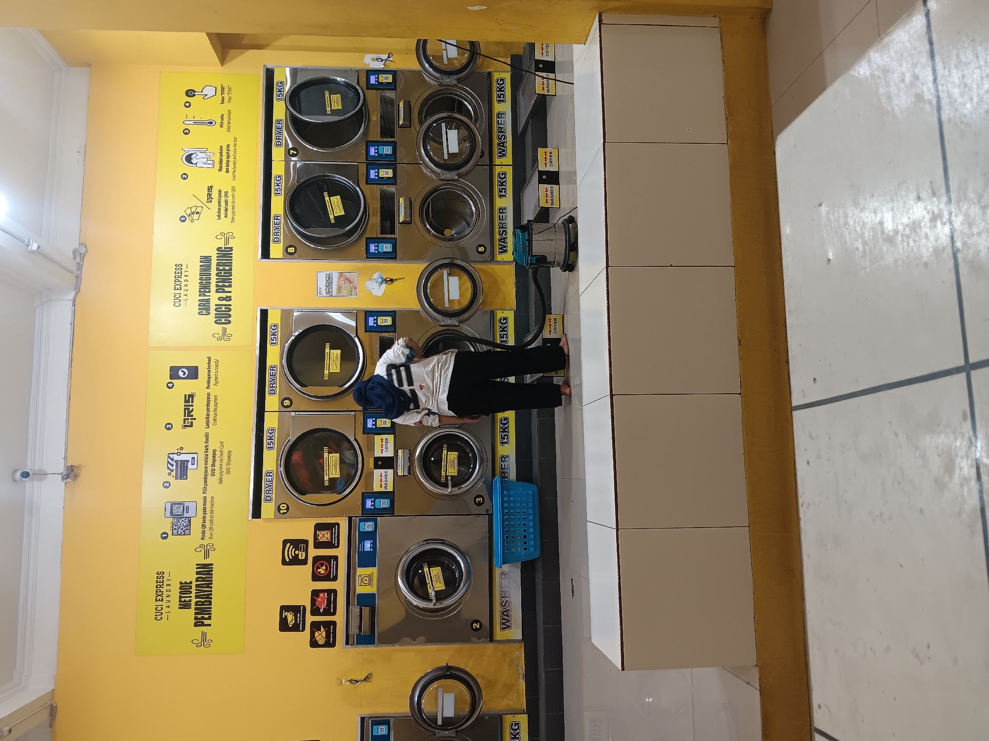 The image depicts the interior of a self-service laundry in Batursari, Indonesia. The walls are painted a bright yellow. A prominent yellow sign on the wall displays instructions for payment methods and how to use the washing and drying machines. The sign has sections titled "CUCI EXPRESS - LAUNDRY - METODE PEMBAYARAN" and "CUCI EXPRESS - CARA PENGGUNAAN CUCI & PENGERING".
There are multiple rows of washing and drying machines. In the foreground, a person wearing a white shirt with dark stripes and black pants, and a blue hijab, is standing in front of a washing machine, appearing to load it. To the left of this person is a blue plastic laundry basket. Several washing machines and dryers are visible, with some labeled as "WASHER 15KG" and "DRYER 15KG". The machines have digital displays and QR codes for payment. A vacuum cleaner is positioned on the floor in front of some of the machines.
The floor is tiled in a white and grey pattern. The environment appears clean and well-lit, with overhead lighting visible in the ceiling. The overall impression is a modern, functional laundry facility.