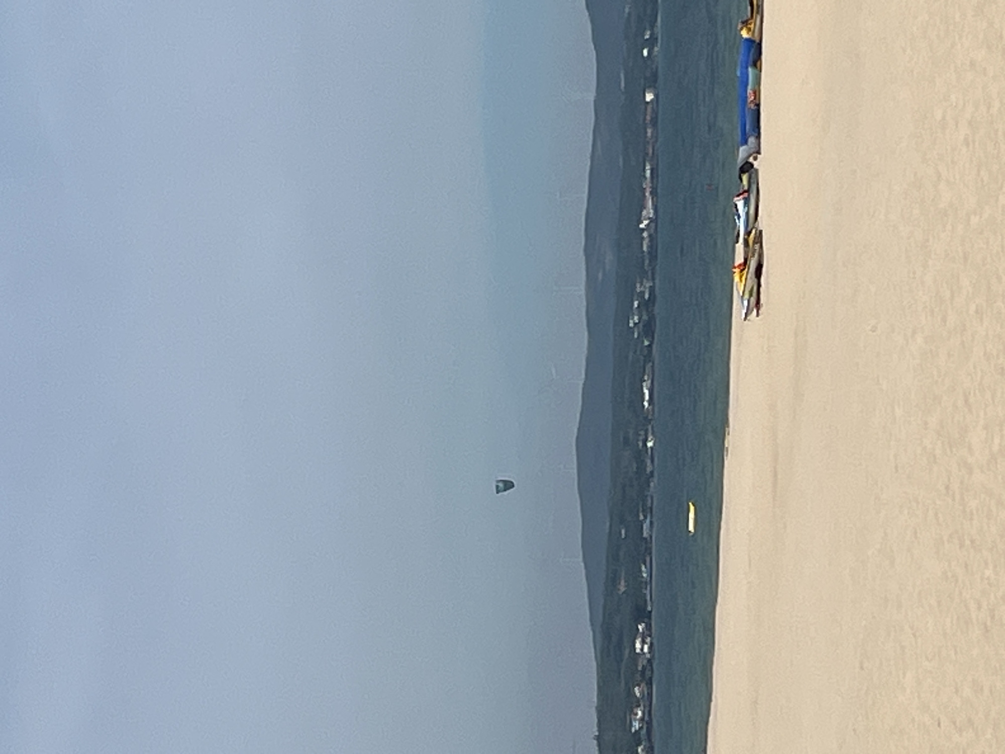 The image depicts a serene beach scene in Phu Thuy Ward, Vietnam, under a hazy, overcast sky. The foreground is dominated by a wide expanse of pale, sandy beach. Along the edge of the beach, several colorful kayaks and surfboards are neatly arranged, suggesting preparations for water activities. In the middle ground, a calm, blue body of water stretches towards a distant, hilly coastline. A small, yellow inflatable object is visible floating on the water. The coastline is dotted with a cluster of buildings, indicative of a coastal town. Further in the distance, several wind turbines stand on the hills, silhouetted against the pale sky. High in the sky, a single kite is captured in mid-air, appearing as a small, dark shape against the muted backdrop. The overall impression is one of a quiet, coastal recreational area.