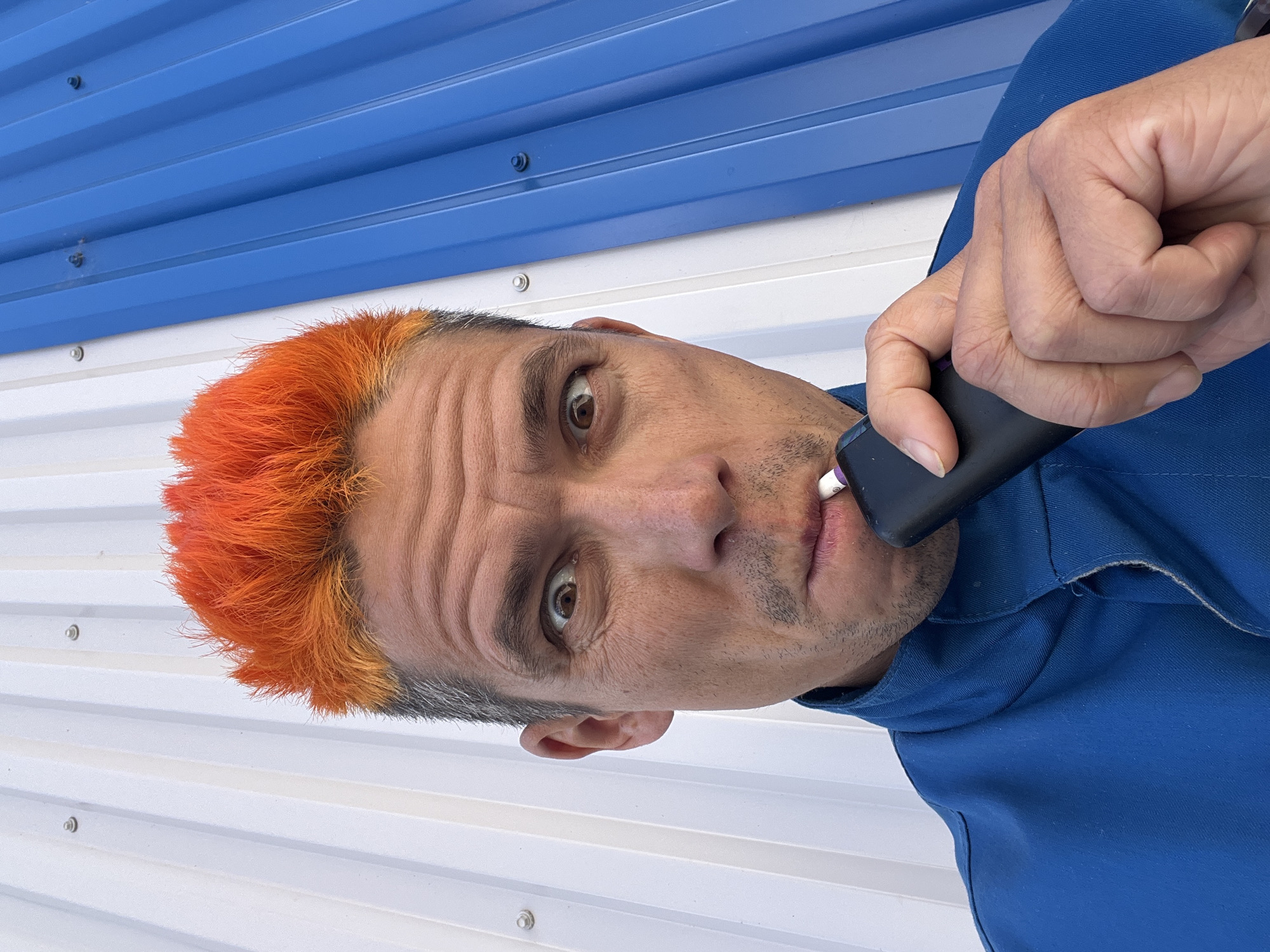A man with short orange hair is holding a black electronic cigarette to his lips. He is wearing a blue shirt. The background is a corrugated metal wall painted white and blue. The location is Chaikovsky, Russia.