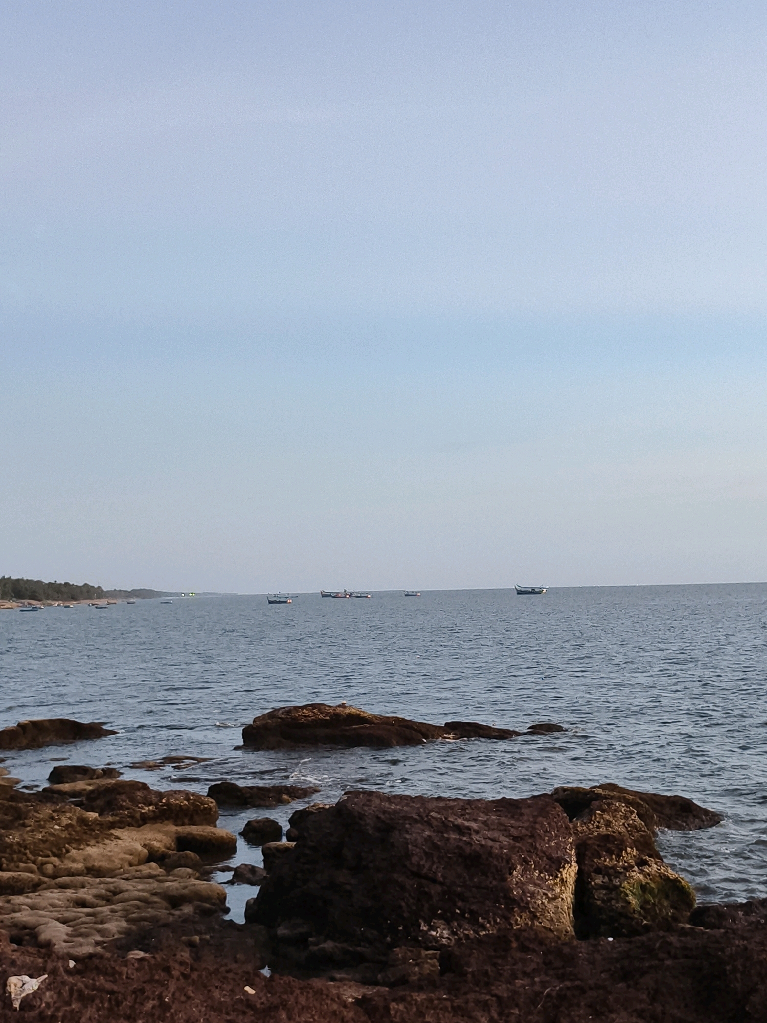 The image depicts a serene coastal scene, likely in Anangadi, India. The foreground is dominated by a rocky shoreline, with rough, textured stones in shades of brown and red. Some of these rocks are partially submerged in the gently lapping water, creating small pools and reflections. The water itself is a calm, deep blue, stretching out to the horizon.
Further out at sea, several small fishing boats are visible. They are scattered across the water, some appearing to be stationary, while others may be in motion. The boats are of a traditional design, common in coastal fishing communities. In the distance, the coastline is lined with trees, creating a soft green border against the blue of the sea and sky. The sky is a pale, clear blue with a hint of haze, suggesting it might be late afternoon or early morning. The overall impression is one of peaceful solitude and natural beauty.