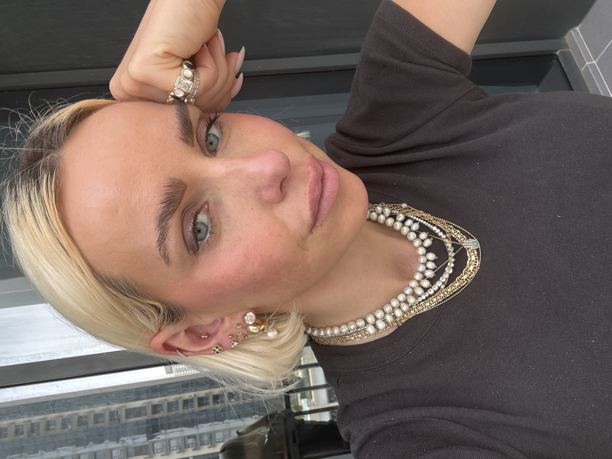 A blonde-haired woman in a dark-colored shirt poses, facing the camera. She is adorned with multiple rings on her left hand and layered necklaces of pearls and gold. Her ears are decorated with numerous earrings. In the background, a window reveals a cityscape, suggesting the location is Dubai, United Arab Emirates. Buildings are visible through the glass.