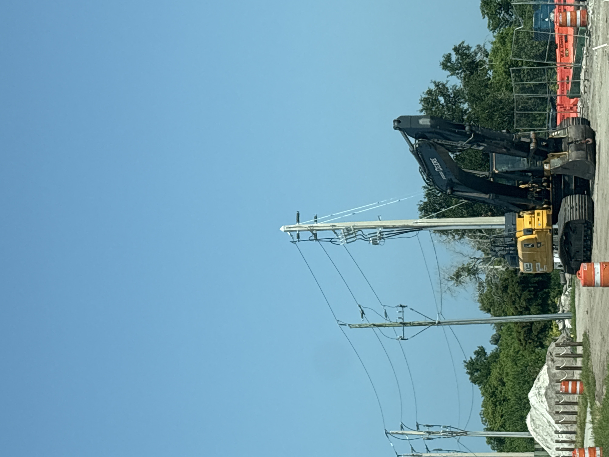 This image, captured in Bradenton, Florida, shows a construction scene under a clear blue sky, suggesting it is daytime with fair weather. The primary subject is a large, dark-colored excavator with "DEERE" visible on its arm, indicating it's a John Deere brand. The excavator is positioned on a gravel surface, and its machinery is extended, implying it is either in operation or ready for use.
In the background, a chain-link fence borders the construction area, with orange traffic barrels and some blue construction barriers visible behind it. Lush green trees form a dense backdrop, suggesting a location with significant vegetation. Also prominent in the foreground are utility poles with multiple power lines stretching across the frame. The lines are suspended on insulators attached to the wooden poles, indicating the presence of active electrical infrastructure.
Further to the right, a large white tarp covers a substantial object, possibly construction materials or equipment. More orange traffic barrels are scattered along the edge of the gravel area. The overall impression is one of ongoing work or preparation for a construction project. There are no people visible in the image.