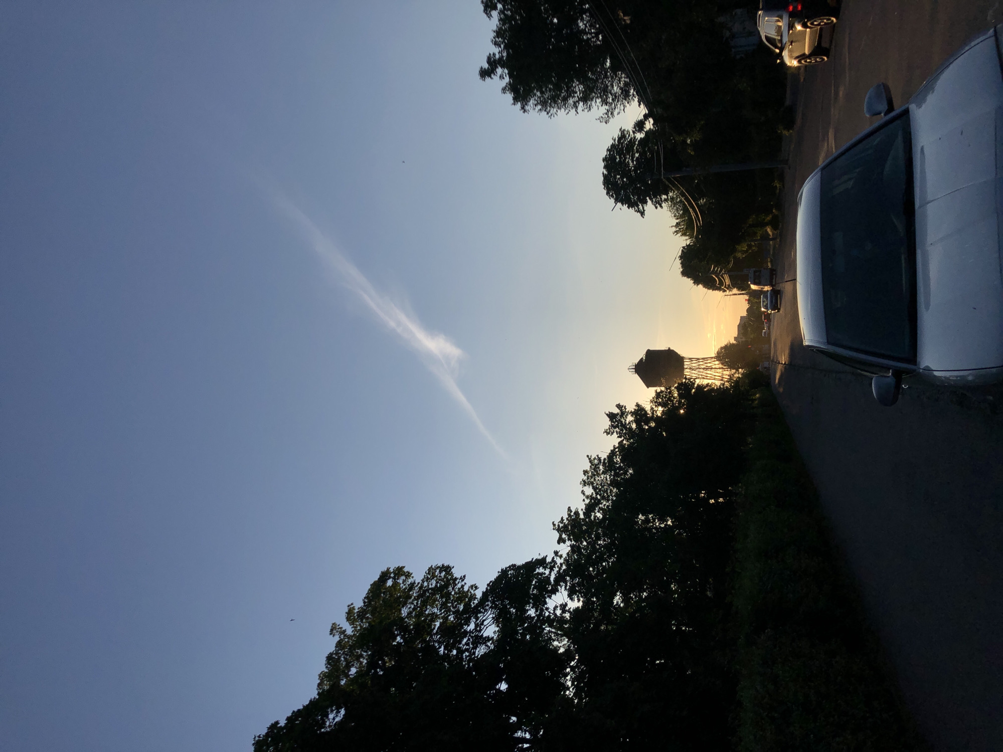 The image captures a serene road scene during late afternoon or early evening, likely at sunset. The sky dominates the upper portion of the frame, transitioning from a clear, deep blue in the upper left to a warm, soft yellow and orange glow closer to the horizon on the right. A single, long, wispy white cloud or contrail stretches diagonally across the sky from the lower left towards the upper right.
Silhouetted against the sky, dark, leafy trees frame both sides of the asphalt road that extends into the distance. On the right side, the hood and windshield of a silver car are prominently visible in the foreground, suggesting the photo was taken from within or beside it. Further down the road, several other vehicles are discernible, including a white car closer to the foreground and another vehicle near the horizon.
A distinctive water tower stands out as a dark silhouette against the glowing horizon line, a common landmark in many towns or suburban areas. The low angle of the sun, casting long shadows and illuminating the sky with warm hues, indicates either sunrise or sunset, with the overall ambiance suggesting the latter. No people or visible text are present in the image. The scene evokes a sense of calm and the natural beauty of twilight.