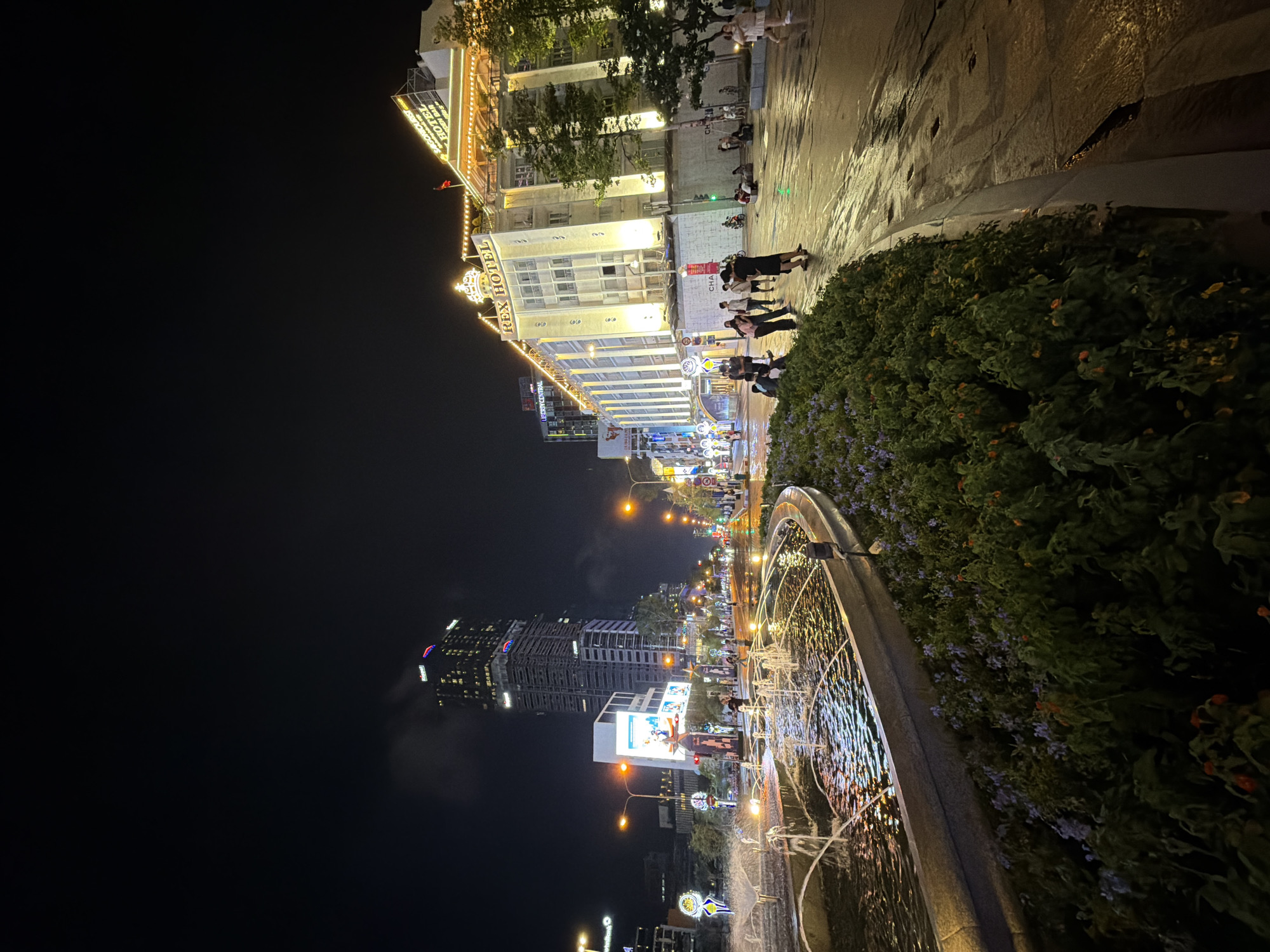 The photograph captures a nighttime cityscape in Thủ Đức, Vietnam. Dominating the foreground is a fountain with water features. Along the walkway, several people can be seen, likely tourists. Illuminated buildings stand in the background, including the "Rex Hotel," identified by its signage. Modern high-rise buildings are also present, adding to the city skyline. Streetlights illuminate the scene, and the overall ambiance is that of an evening setting.
