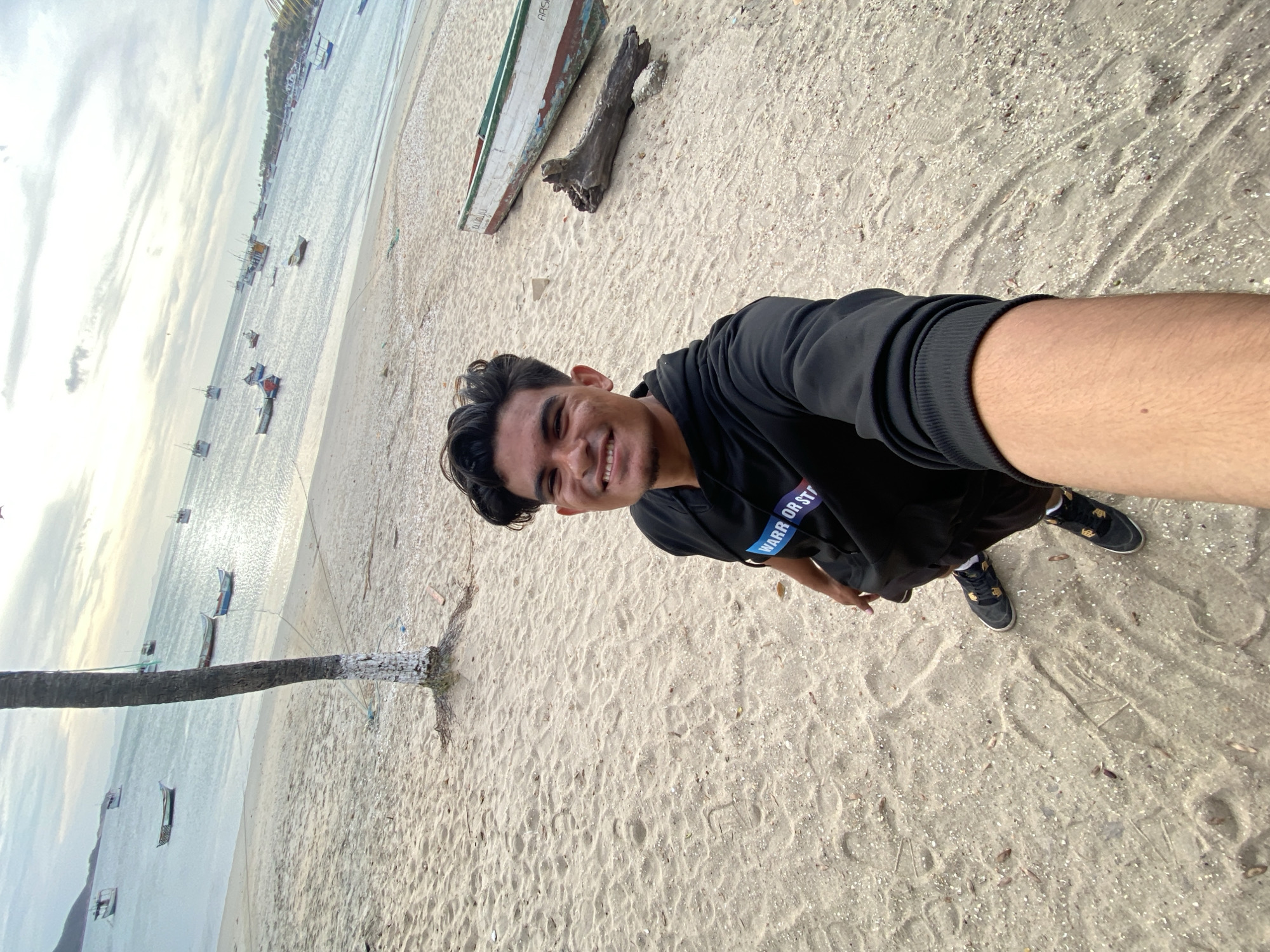 An image taken on a sandy beach in Juan Griego, Venezuela, shows a young male subject in a selfie-style shot. The subject, positioned centrally, is a male with dark hair, dark eyes, and a visible mole on his left cheek, smiling directly at the camera. He is wearing a black hoodie with a partial blue and white graphic reading "WARR OR ST" on the chest, dark shorts, and black sneakers with white soles and gold detailing. His right arm is extended, holding the camera.
The foreground consists of light-colored sand with scattered small debris and footprints. To the subject's left, a palm tree trunk, painted white at its base, extends upwards from the sand. To the subject's right, further back on the sand, lies a weathered wooden boat, painted white with a green trim, and a large piece of driftwood.
The background features a body of water, likely the Caribbean Sea, with numerous small fishing boats, many with masts, scattered across its surface. The water is calm and appears light blue-green. In the distant background, hills are visible on the horizon. The sky is overcast with scattered clouds, indicating diffused daylight.
