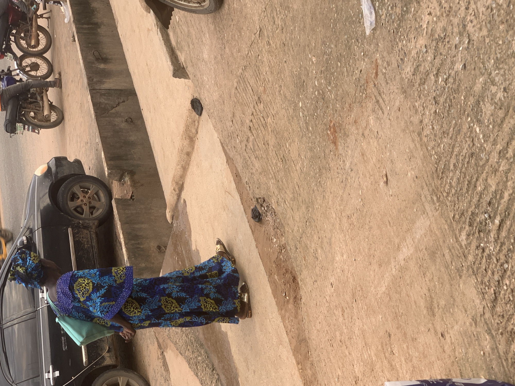 In Shagamu, Nigeria, a person wearing a blue and yellow patterned traditional dress and a matching headwrap stands near a black car and two motorcycles on a dirt and concrete surface. The person carries a light blue bag over their left shoulder and wears gold sandals. The environment appears to be a roadside area with a concrete drainage ditch. The person's back is to the camera, and they are looking away from it. One of the motorcycles is parked in front of a person wearing jeans and brown sandals, with their legs visible.