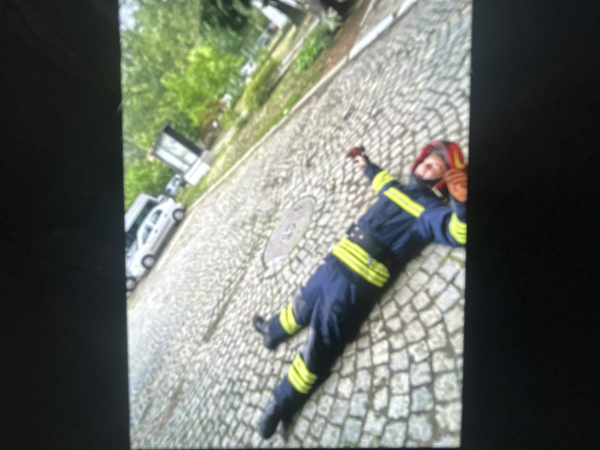 A firefighter in full uniform, including a helmet, is lying prone on a cobblestone street in a city, presumed to be Unknown, Unknown. The street also has a manhole cover. A white vehicle, possibly a van, is parked nearby. Trees are visible in the background.