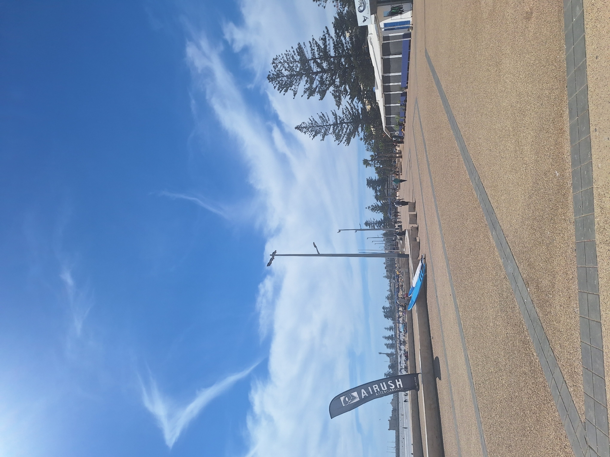 This image depicts a sunny day at a promenade in Essaouira, Morocco, likely along the coastline. The foreground is dominated by a textured, sandy-colored pavement with grey rectangular inlays. To the left, a black flag with white lettering, reading "AIRUSH KITEBOARDING," stands prominently. Nearby, a blue surfboard and a white object, possibly another board or accessory, rest on a slightly elevated concrete ledge.
In the midground, a wide promenade stretches out, populated by a few pedestrians walking in both directions. Along the right side of the promenade, there is a building with a white exterior and blue trim, possibly a cafe or shop, with outdoor seating. Several tall, slender pine trees are visible behind the building. Further in the distance, along the coastline, are more trees, buildings, and a sandy beach with numerous people enjoying the seaside. The ocean is visible in the far left of the image, with a faint outline of a historical building or fortification on a hill. The sky is a vibrant blue, punctuated by wispy white clouds, suggesting fair weather.
