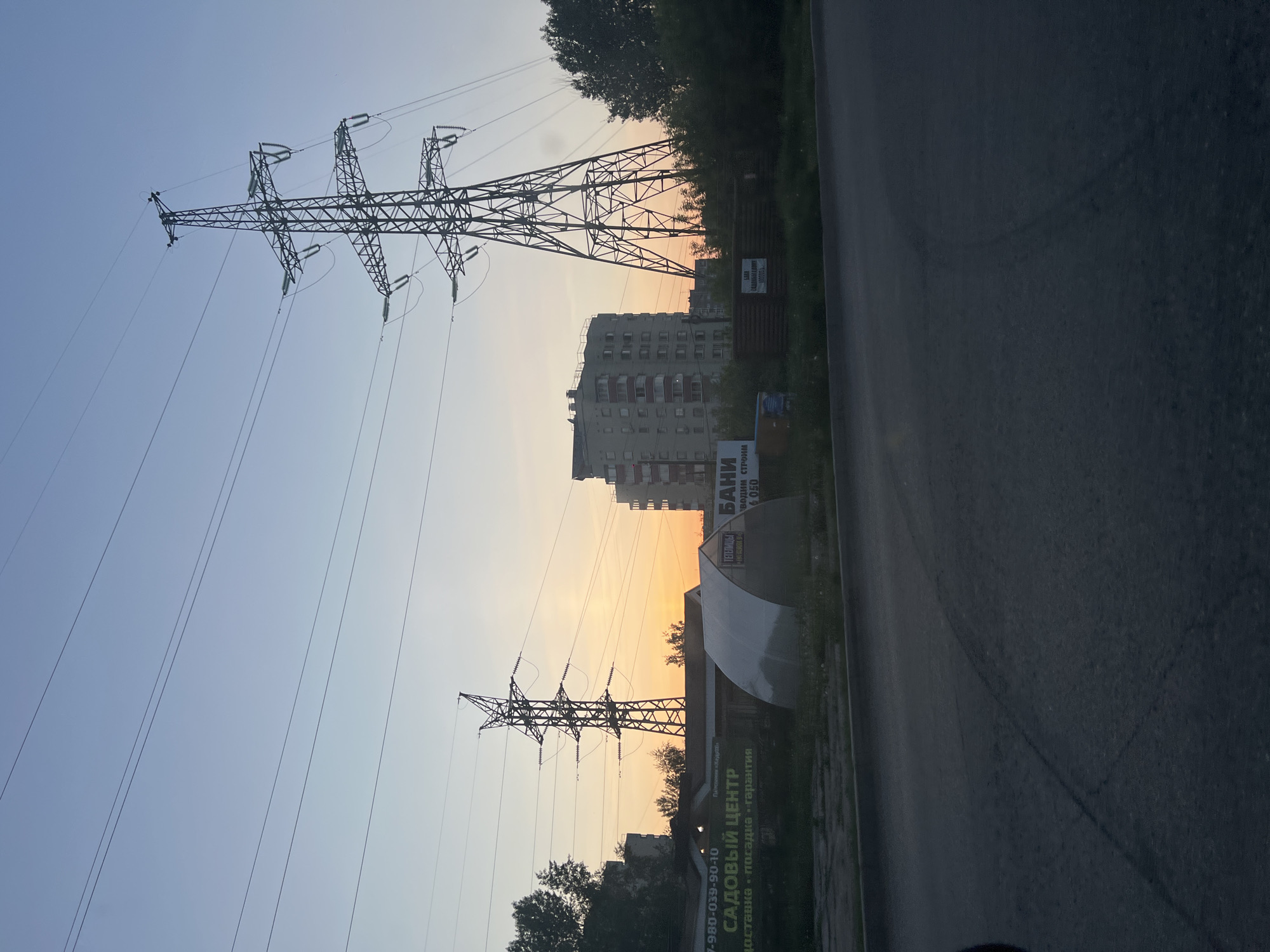 The image shows a street scene in what appears to be a residential area of Chaikovsky, Russia. In the foreground are two tall electrical transmission towers with multiple power lines extending across the sky. Behind the towers, a multi-story apartment building is visible against a sky that transitions from light blue to a warm, orangish hue near the horizon, suggesting either sunrise or sunset. A commercial sign is present on the right side of the frame and part of a billboard is seen. The ground level along the road is covered with vegetation. The road itself is dark and occupies the right third of the frame.