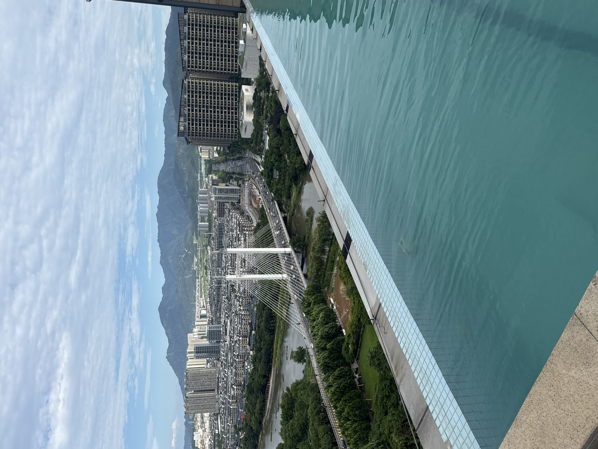 This is an aerial shot of the city of Yiwu, China, taken from a rooftop infinity pool. The pool, with its turquoise water and tiled edge, occupies the foreground of the image, leading the eye towards a panoramic view of the city. Beyond the pool, a river winds its way through the urban landscape, spanned by a modern suspension bridge carrying traffic. Numerous high-rise buildings, both residential and commercial, dominate the cityscape, interspersed with green spaces and trees. In the distance, a range of hazy, tree-covered mountains forms a natural backdrop to the sprawling city. The sky is filled with a mix of white, puffy clouds against a pale blue expanse, suggesting fair weather. No people are visible in the frame.
