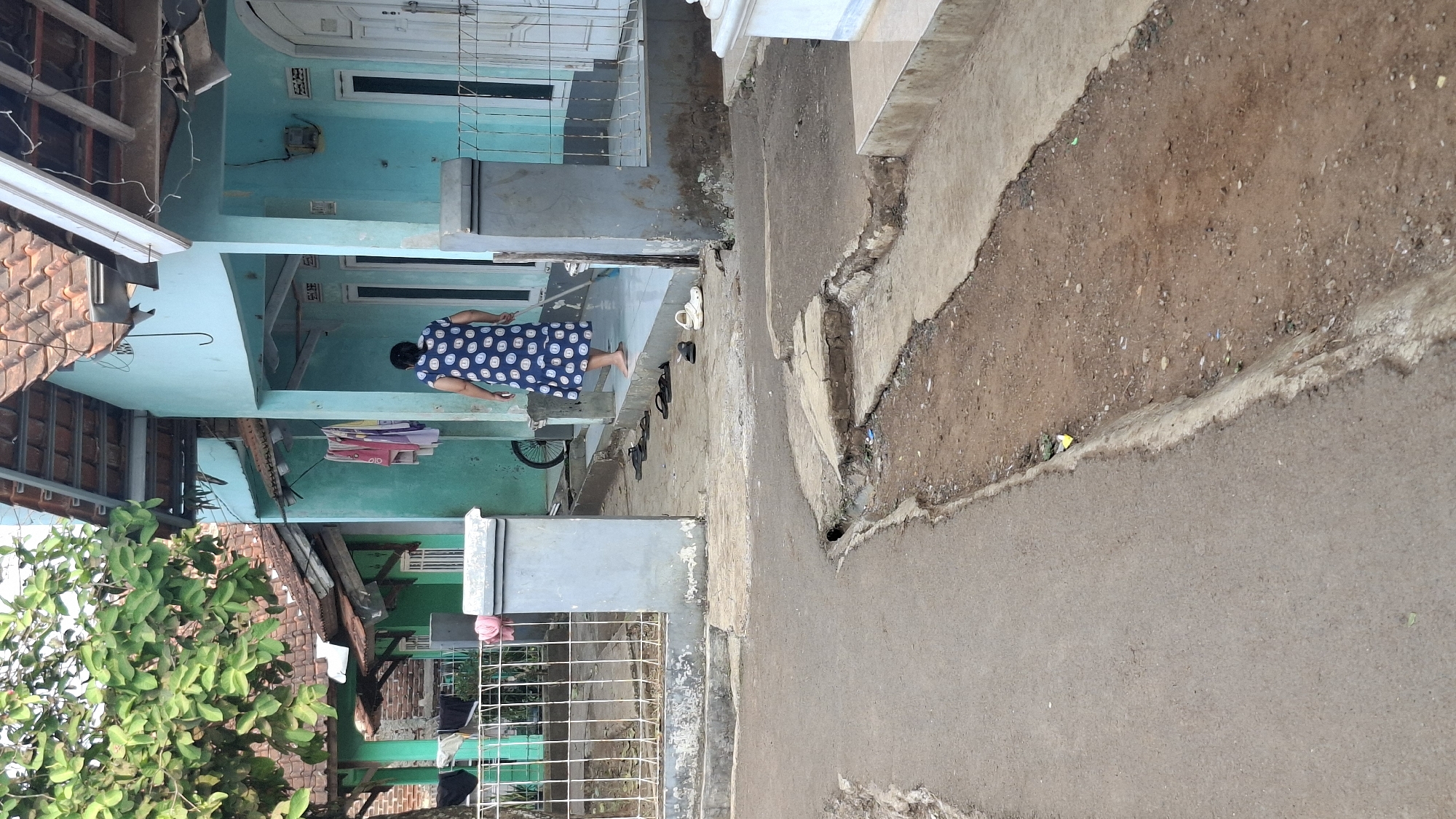 An elderly woman wearing a blue and white polka dot dress is sweeping the porch of a light blue house. She is barefoot. Several pairs of sandals are scattered on the porch and in front of the house. A metal gate with a white concrete base separates the property from a dirt and asphalt road. The road has a drainage ditch running along its edge. Clothes are hanging on a line in front of the house. Trees with green leaves are visible. The house has a red tile roof. The scene appears to be in a residential area in Cilaku, Indonesia.
