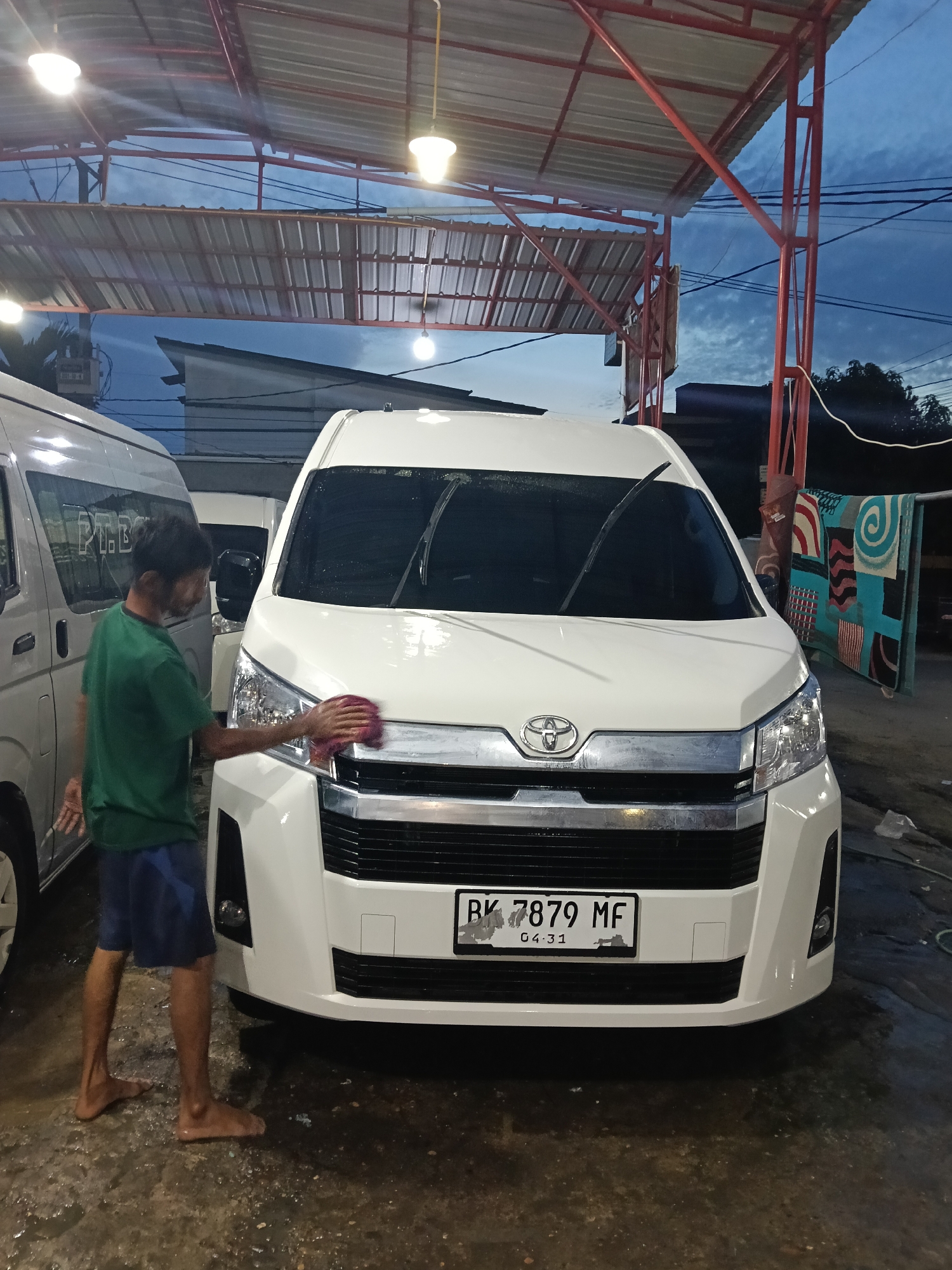 In Lhokseumawe, Indonesia, a man in a green t-shirt and blue shorts is washing a white Toyota HiAce van with a red cloth. The van has license plate "BK 7879 MF" and is parked in a covered car wash area with a metal roof structure supported by red metal beams. The floor is wet. Another van is parked to the left, partially visible. In the background, a building and part of the twilight sky are visible. To the right, colorful patterned cloths hang on a rack.