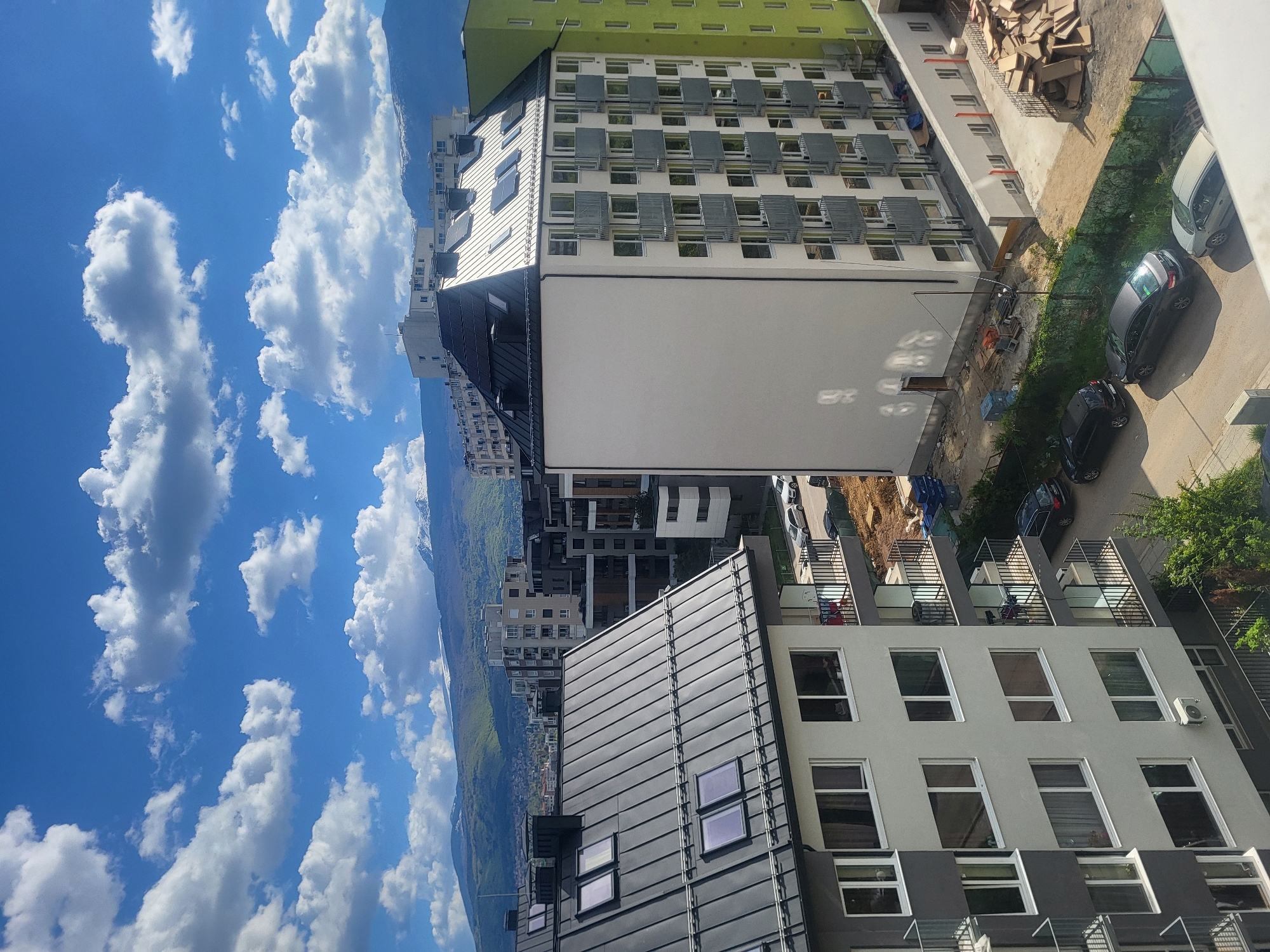 The image shows an elevated view of buildings and a street scene in Sofia, Bulgaria. The sky is blue with puffy white clouds. Buildings of varying heights and architectural styles are present, some with green accents. Cars are parked on the street, and construction materials are visible on a rooftop. Mountains are visible in the distance.