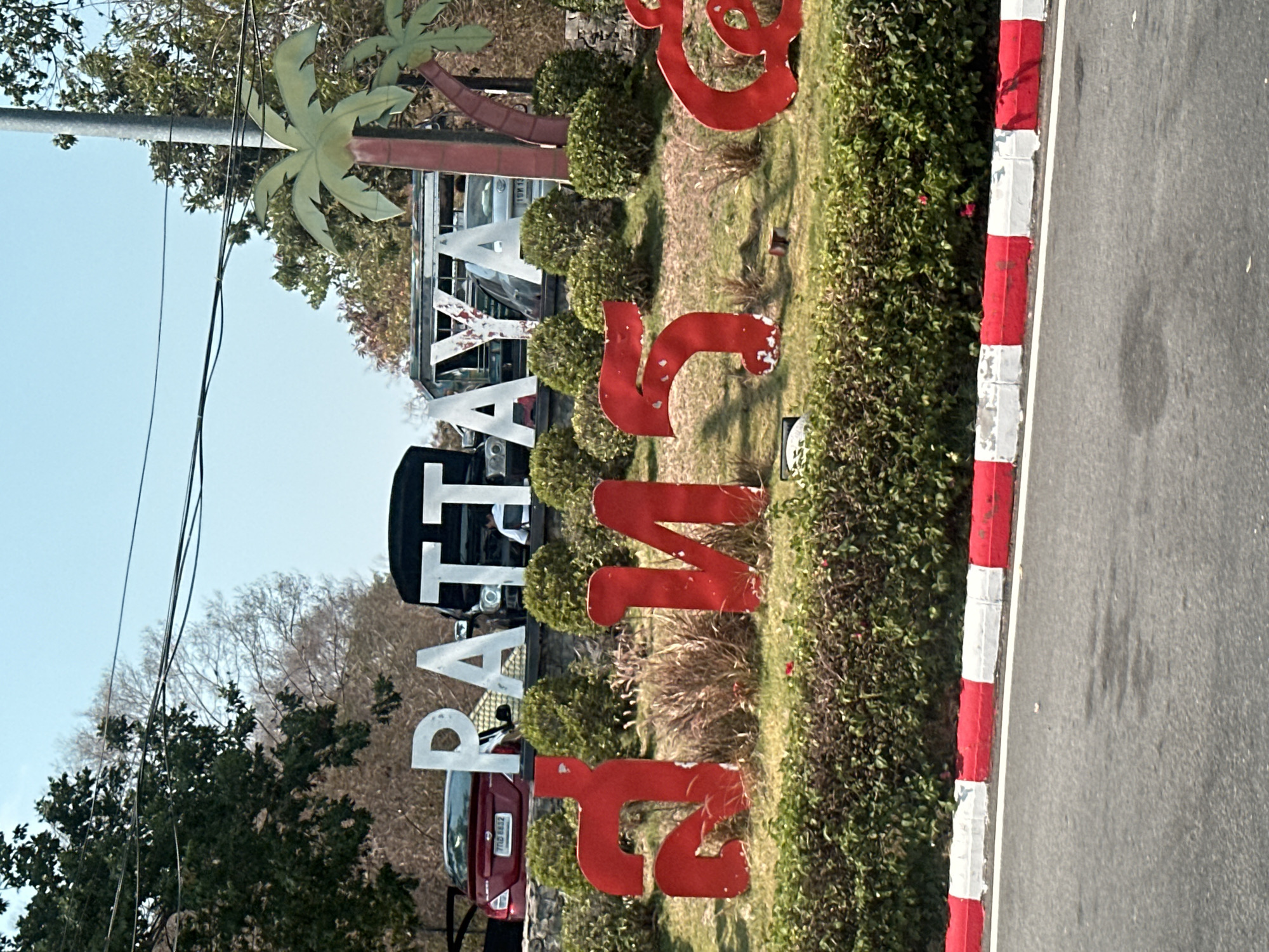 The image captures a sign reading "PATTAYA CITY" with "พัทยา" beneath it. The letters of "PATTAYA" are white, while the Thai script is in red. The sign is situated in a grassy area with surrounding bushes. Cars are parked behind the sign, suggesting its placement along a roadside. A red and white curb marks the edge of a paved road to the right. Overhead, a utility pole with wires is visible against a clear sky.