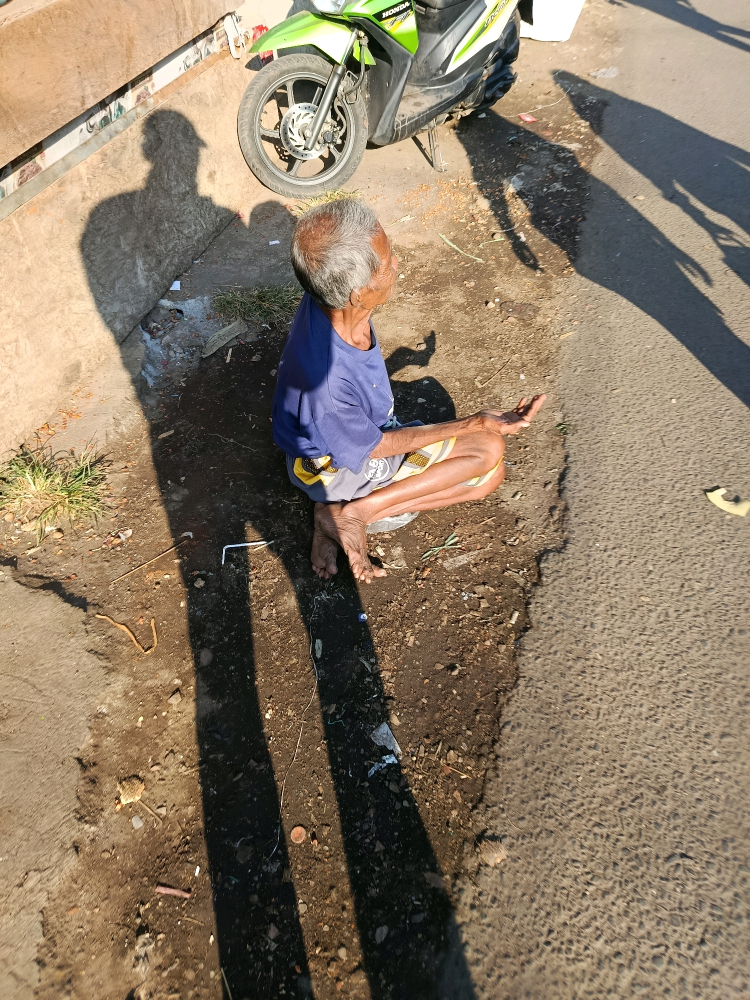 An elderly individual with graying hair is seated on the ground, facing away from the viewer, in what appears to be a roadside location in Gondanglegi, Indonesia. The person is wearing a blue t-shirt and patterned shorts. Their left hand is extended, palm up, in a gesture that could be interpreted as asking for something. The ground is a mix of dirt, gravel, and some debris. A portion of a light green and white scooter is visible in the upper left quadrant of the image. Long shadows are cast across the scene, indicating it is likely late afternoon or early morning. The overall environment suggests an outdoor, public space, possibly near a street or pathway.