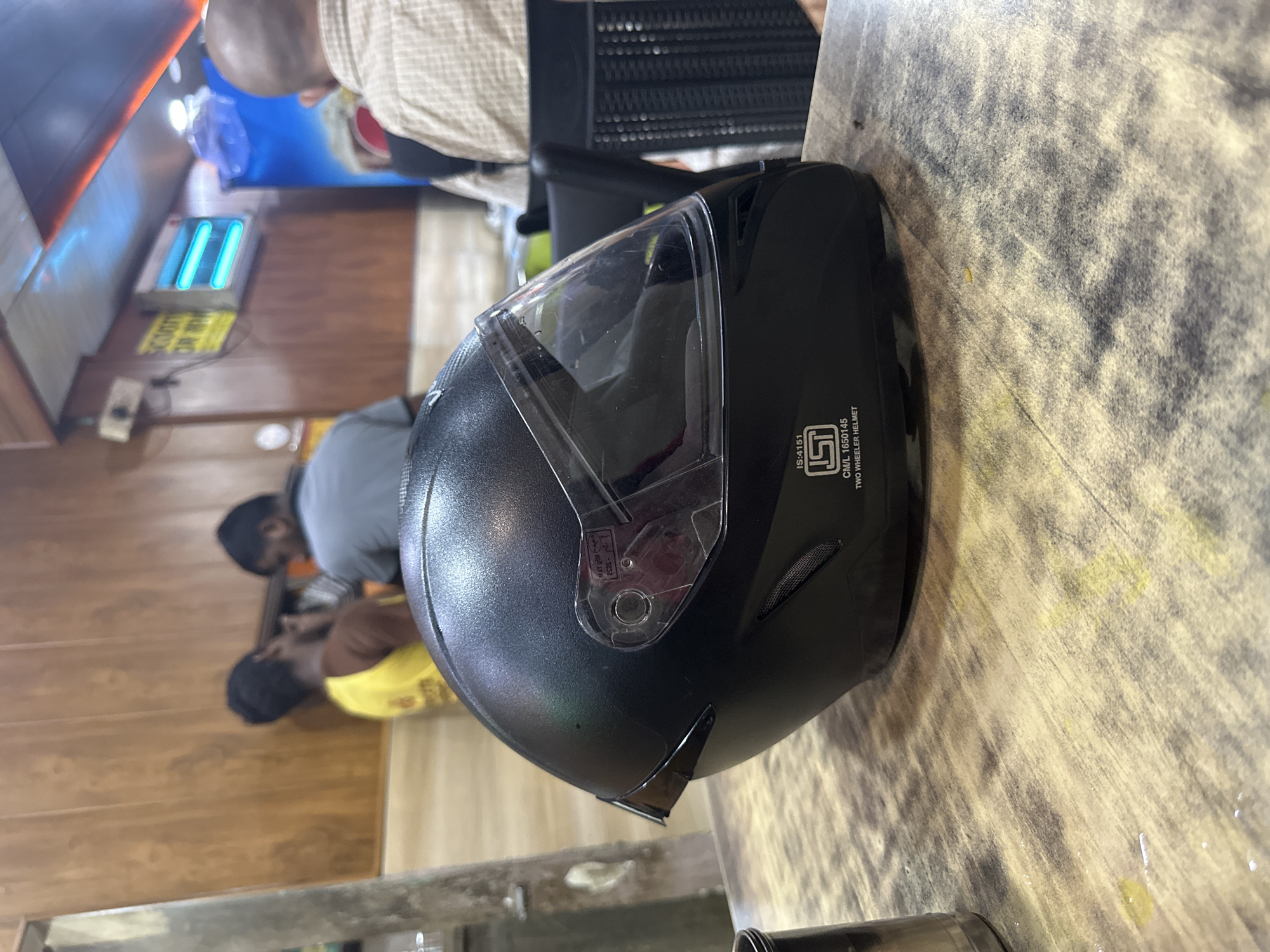 A black motorcycle helmet with a clear visor sits on a wooden table in the foreground. The helmet has an "ISI" certification sticker on its side, indicating it is a two-wheeler helmet. In the background, two men are visible, one wearing a yellow shirt and the other a gray shirt. A third person, seen from behind and wearing a checkered shirt, is seated at a table to the right. The environment appears to be an indoor establishment with wooden paneling on the walls and fluorescent lights on the ceiling.