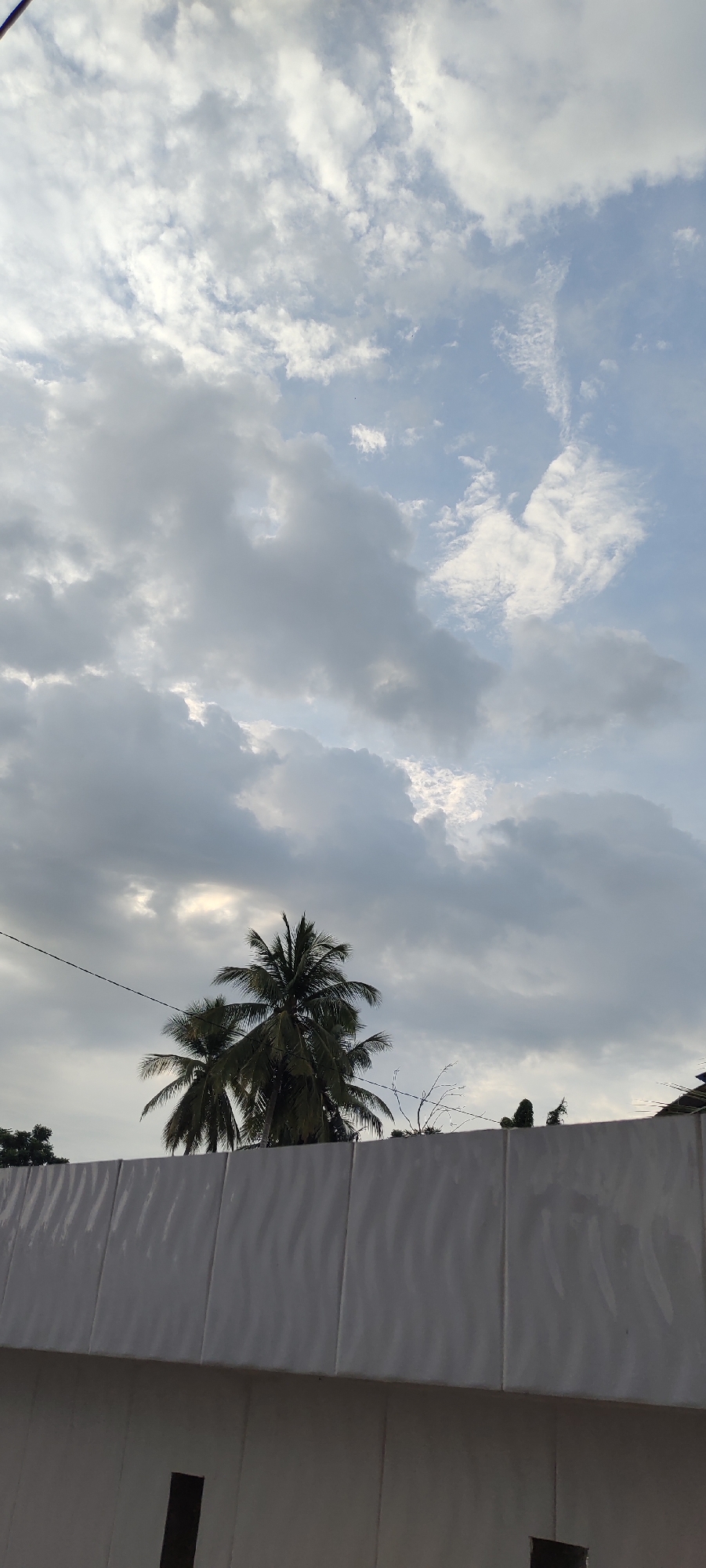 The image is a vertical, outdoor shot taken at ground level, looking upwards. The foreground is dominated by a white, textured wall with visible seams running vertically. Above the wall, two coconut trees with lush green fronds are silhouetted against a cloudy sky. The sky is a mix of light blue and grey clouds, with patches of brighter sky visible through breaks in the clouds. A thin black wire stretches horizontally across the frame above the trees. The scene appears to be a view of the sky and vegetation from behind a wall, possibly in a residential or urban area of Meunasahaluedrienlhoksukon Barat, Indonesia. No people are visible in the frame.