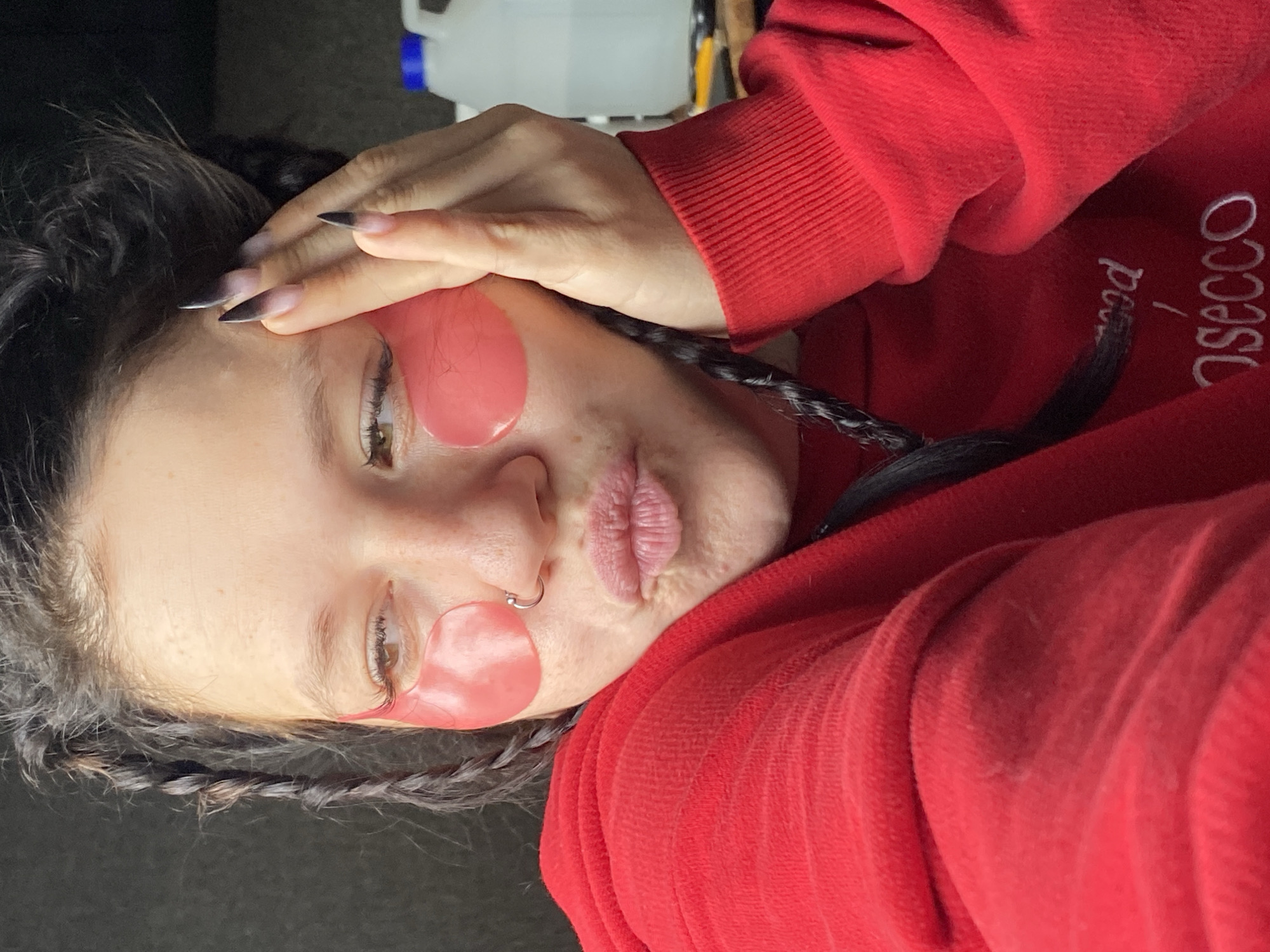 A close-up, selfie-style shot shows a young woman with dark hair styled in braids, wearing pink under-eye patches and a bright red hoodie. Her left hand is raised, with long, dark, pointed nails touching her face near her right eye. She is looking to her left with her lips pursed. A silver nose ring is visible. The background is out of focus, but appears to be indoors with some bottles and objects on a shelf visible in the upper right corner. The overall impression is casual and personal, likely taken in a private setting. The image does not contain any specific visual cues that would allow for the determination of its location in Chaikovsky, Russia.