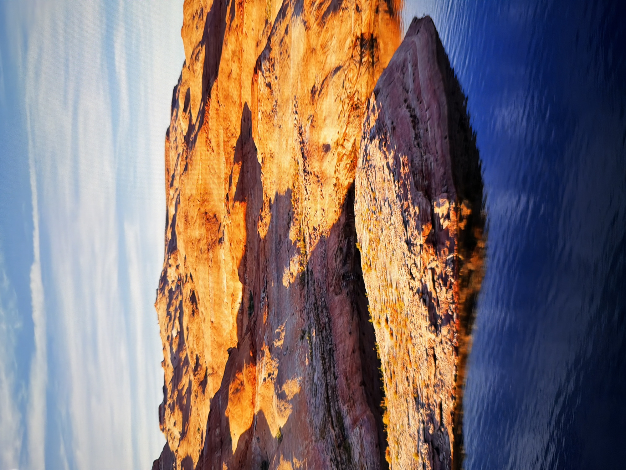 This is a vertical, high-angle, close-up photograph of a desert landscape with a body of water in the foreground. The sky is blue with wispy white clouds, and the sun appears to be low on the horizon, casting warm, orange light on the desert formations. The desert is characterized by rocky, layered formations with deep shadows. A small, rocky island is situated in the body of water. The water is dark blue and reflects the colors of the sky and the desert. The scene appears to be a natural, undeveloped area.
This image does not depict the city of Kyiv, Ukraine.