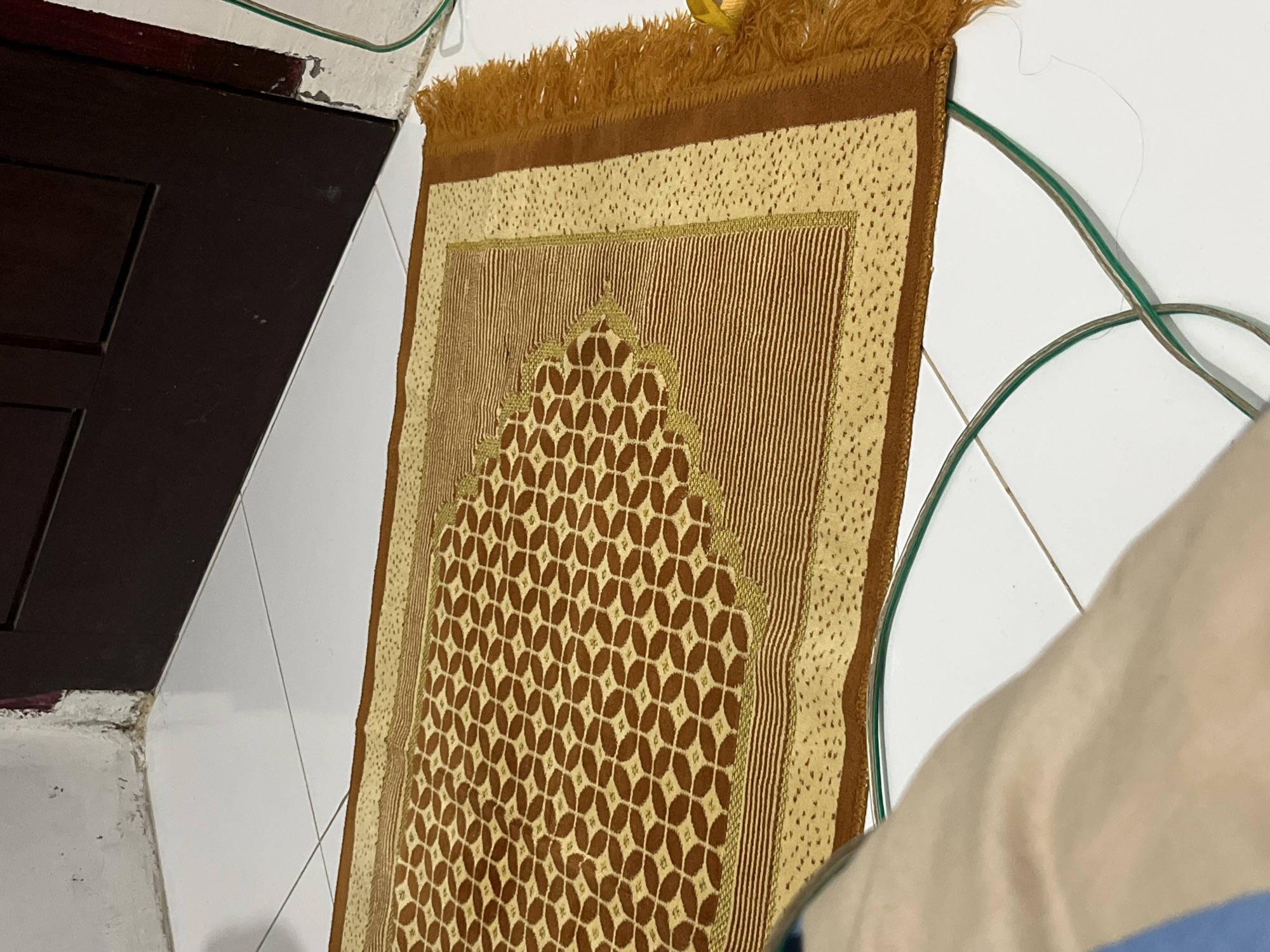 This image presents an indoor scene, likely within a residence in Lhokseumawe, Indonesia. The central subject is a prayer mat (sajadah), predominantly brown and light beige or cream, laid against a white tiled wall. The mat is intricately patterned with geometric designs, featuring a prominent arch-like motif, characteristic of a mihrab, at one end. Fringes are visible along the top edge of the mat.
To the left, a portion of dark brown wooden furniture, possibly a cupboard or door, is visible, showing paneling details and resting against a white wall with visible tile lines. The base of this wall shows some wear or damage. Green electrical wires are loosely arranged against the white tiled wall, running across the upper right and central right sections of the frame. In the immediate foreground on the right, a blurry light-colored fabric, resembling a pillow or part of bedding, partially enters the frame.
No people are present, and no specific activity is captured, though the presence and design of the prayer mat strongly suggest its use for Muslim prayers. The lighting indicates an indoor setting, but the time of day or weather cannot be determined. There is no visible text in the image. The overall setting appears to be a functional, lived-in interior space.