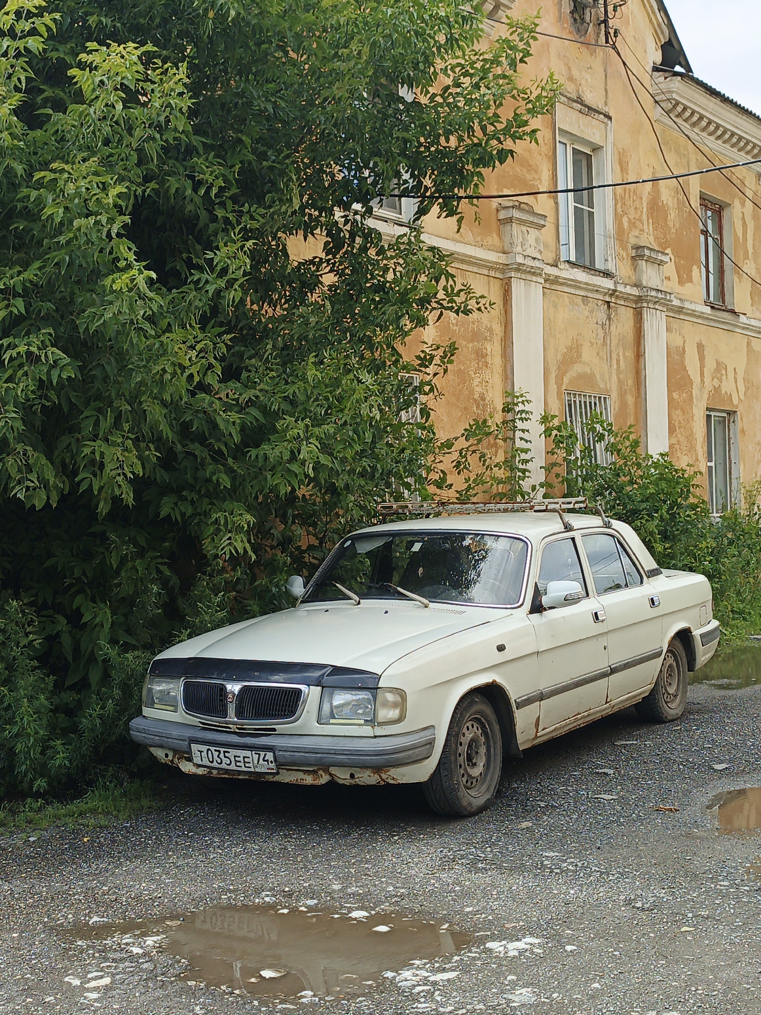 An aged, cream-colored sedan, bearing Russian license plate "T035EE 74", is parked on a gravel driveway next to a dense, overgrown green bush. The car, showing signs of rust on its lower panels and bumper, has a roof rack fitted. Behind the bush and car stands a dilapidated, two-story building with a yellow, weathered facade and numerous windows, some with barred openings. The sky above is overcast. Puddles of water are visible on the ground in front of and to the side of the car, indicating recent rainfall. This scene is located in the city of Zlatoust, Russia.