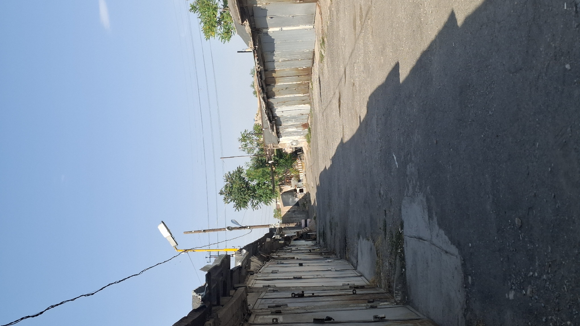 The image depicts a street lined with a row of garages on the left and another row on the right, creating a narrow, paved alleyway. The garages appear to be made of metal, with individual roll-up doors. Some of the garage doors are closed, while others are partially open. The scene is outdoors, under a clear blue sky. Vegetation, including trees, is visible in the background, behind the garages on the left. A yellow lamppost with a modern light fixture stands near the beginning of the alley on the left. Power lines are strung overhead. The ground is paved with asphalt and shows signs of wear, with some patches of dirt and debris. The overall impression is of a utilitarian area, possibly a garage complex or an industrial yard. The image is taken from a low angle, looking down the alleyway. It is daytime, with shadows cast by the garages, indicating the sun is positioned to the right of the frame. There are no people visible in the image.