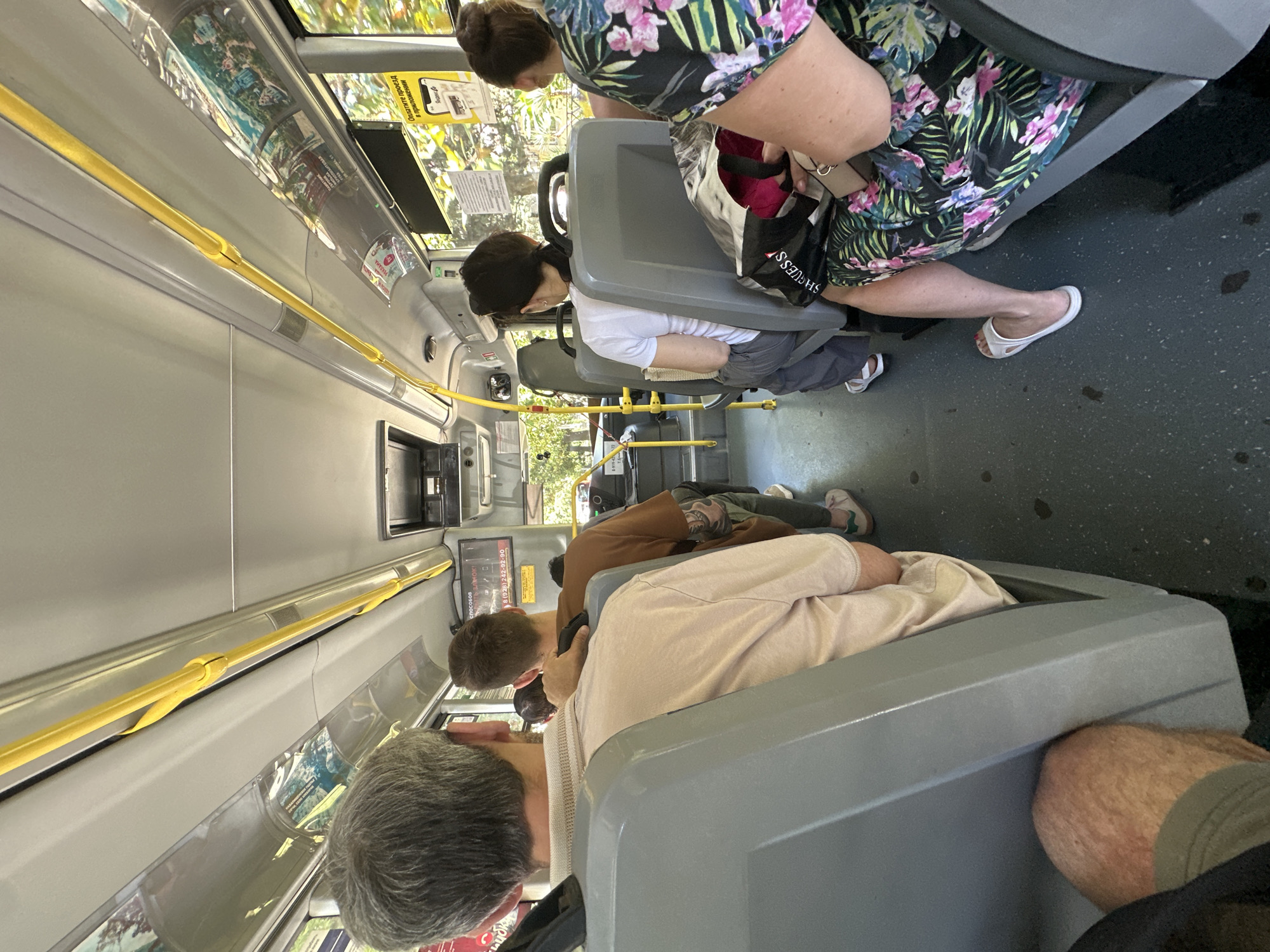The image depicts the interior of a bus with several passengers seated. The bus appears to be in motion, with sunlight streaming in through the windows, suggesting it's daytime. The setting is a public transportation vehicle, likely a bus, as indicated by the rows of seats, overhead handrails, and interior design.
The subjects are primarily passengers. In the foreground, a man with graying hair is seated, facing forward. Behind him, other passengers are visible, some looking towards the front and others appear to be resting or looking away. One woman in the middle of the frame is wearing a floral-patterned dress and white sandals. Another passenger is wearing a light-colored t-shirt and appears to be resting their head against a seat. A person in a brown jacket is also visible further back.
The activity is passive – passengers are traveling on the bus. There are no overt actions or events taking place beyond the journey itself.
Notable details include the yellow handrails running along the length of the bus, a common safety feature. The seats are a muted gray plastic. The windows offer glimpses of greenery outside, indicating a possibly temperate climate. Some advertisements or informational posters are visible on the walls of the bus. There is some visible text on a yellow sign and on black signage above a window, but it is not clearly legible in the provided image. The overall mood is one of quiet travel. The location context of Sochi, Russia, is provided, but there are no specific visual cues within the image that directly confirm this location.